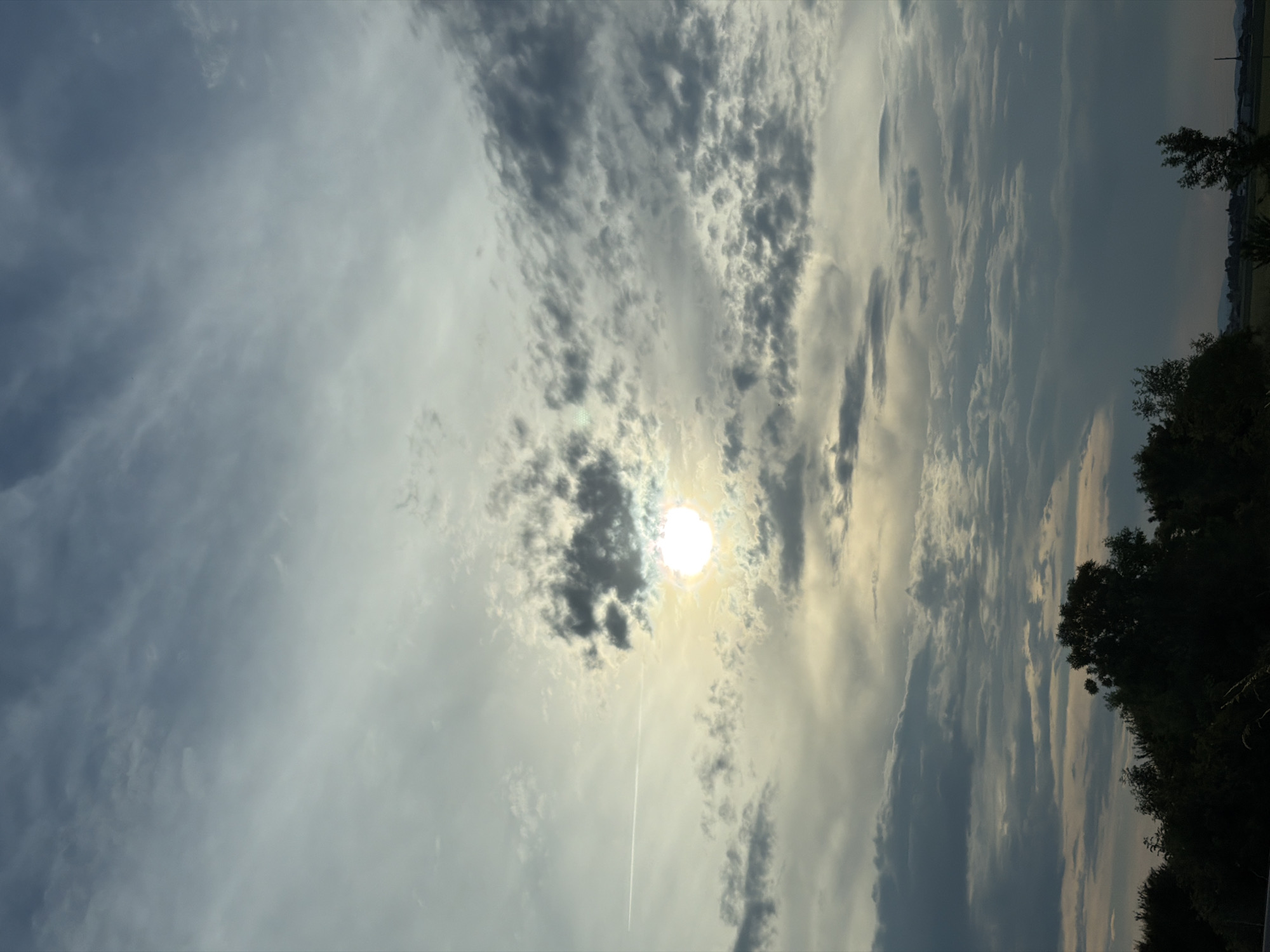 The image depicts a cloudy sky over a landscape, likely in or near Mramor, Bulgaria. The sun is partially obscured by clouds, creating a diffused light that illuminates the sky. A faint contrail from an aircraft stretches across the upper left portion of the frame. The lower part of the image shows a silhouetted treeline against the lighter sky, with hints of distant fields and structures. The overall mood is somber, with the dark clouds and limited sunlight suggesting an overcast day, possibly nearing dusk. There are no people or discernible actions taking place.