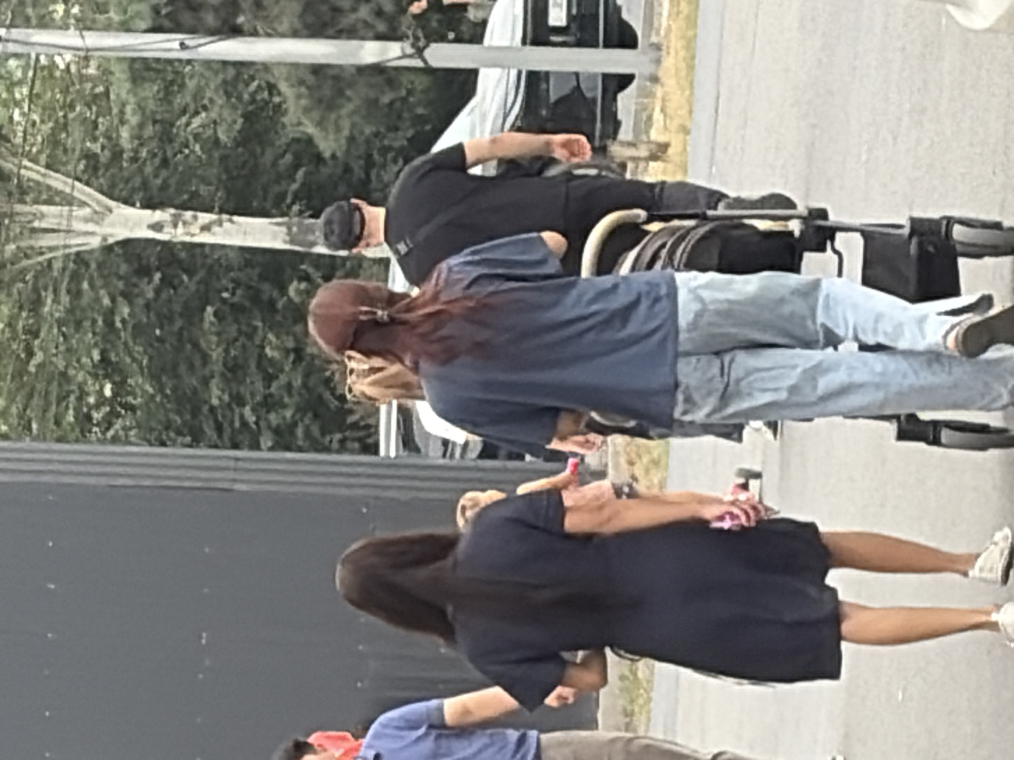 A group of people walk along a street in an urban environment. The foreground features a woman in a dark blue dress and white sneakers, with her back to the camera. Beside her, a person in a blue shirt and light-colored pants also walks away. To the right, a person in a dark blue t-shirt and jeans pushes a stroller. Behind them, a man in a black t-shirt and black pants, wearing a black baseball cap, walks in the same direction. A child in a pink and white outfit is visible between the woman in the blue dress and the stroller. In the background, a dark-colored SUV is parked on the right side of the street, with trees and foliage visible beyond it. The scene takes place on an asphalt surface.