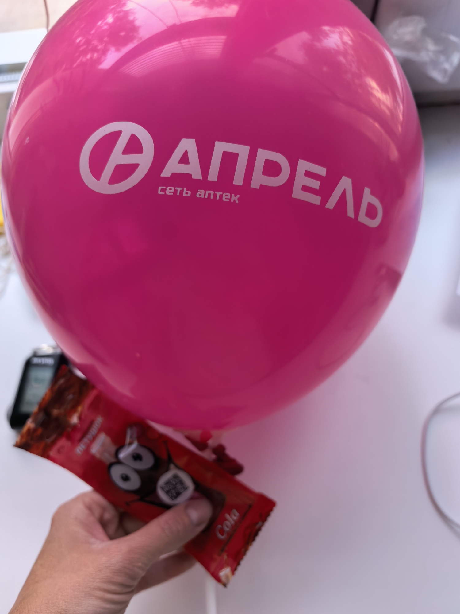 A bright pink balloon with the logo of the Russian pharmacy chain "Апрель" ("April") in white text is the primary subject of the image. The balloon appears to be indoors, likely near a window, given the natural light and partial view of an exterior scene in the reflection. The text "сеть аптек" (pharmacy chain) is written below the main logo.
A hand holds a cola-flavored candy bar, partially obscuring the bottom of the balloon. The wrapper is red with a cartoonish depiction of a character.
Other objects visible include a black electronic device partially visible on the left side, and a white object, likely a wall or windowsill.
Given the "Апрель" pharmacy chain's presence and the location in Russia inferred from the text, it's possible the image was taken in Sterlitamak, Russia. However, without specific identifiers in the image itself, this location is speculative.