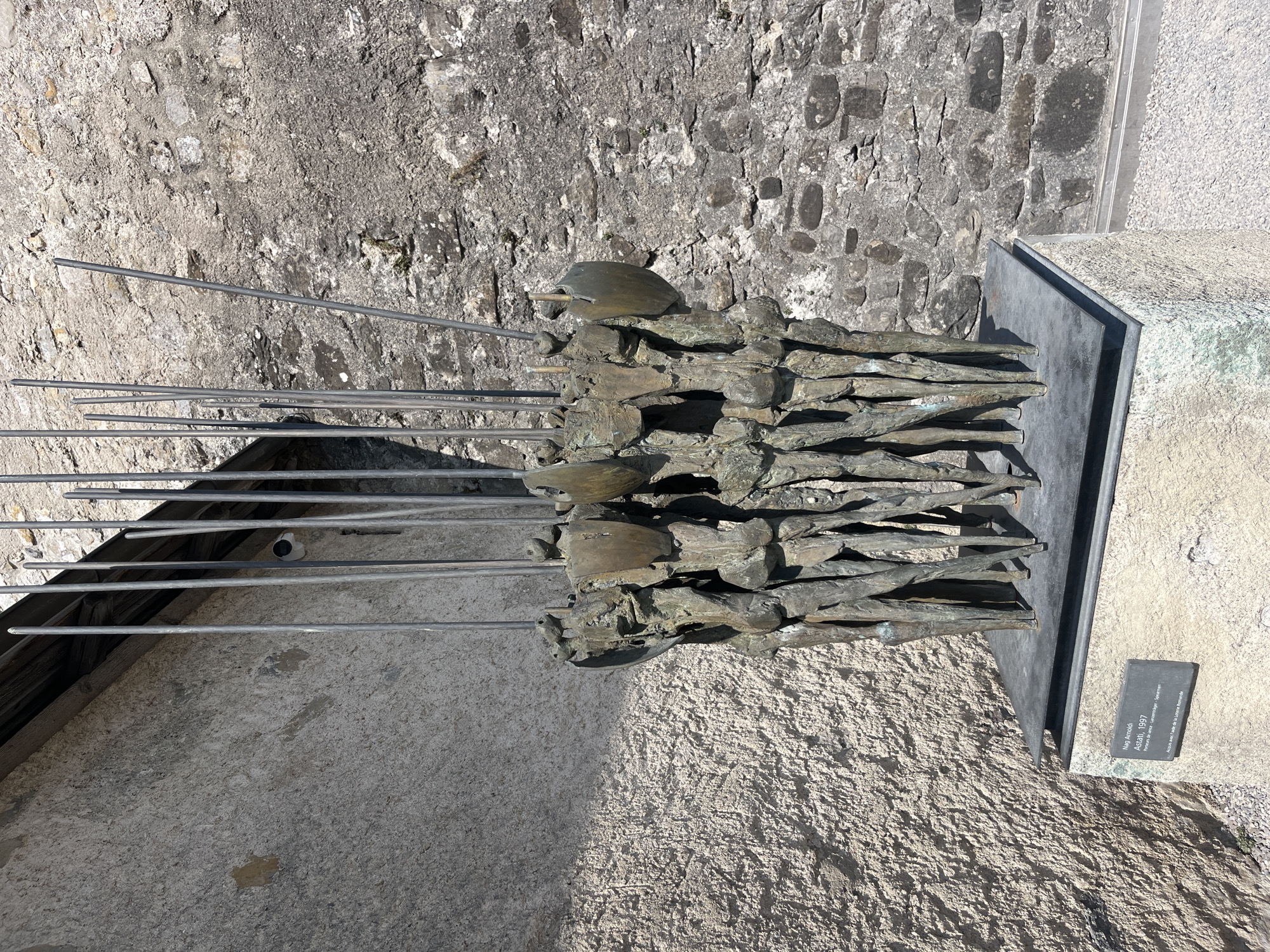 The image shows a bronze sculpture of a group of figures with spears, placed on a stone pedestal against a textured stone wall. A plaque at the base of the pedestal reads "Nag Arnoldi Astati, 1997". The sculpture is in a public outdoor space, as indicated by the stone wall and pedestal. The background wall is constructed of rough-hewn stones, with some areas of plaster or stucco. The sculpture itself consists of multiple figures, tightly grouped together, each with elongated, abstract forms and holding upright spears. Some of the figures appear to have shield-like elements attached to their bodies. The overall impression is one of a unified, perhaps martial, formation. The environment appears to be sunny, casting shadows on the sculpture and the wall. The location is identified as Epagny, Switzerland, though the exact placement within the city is not specified by the visual information alone.