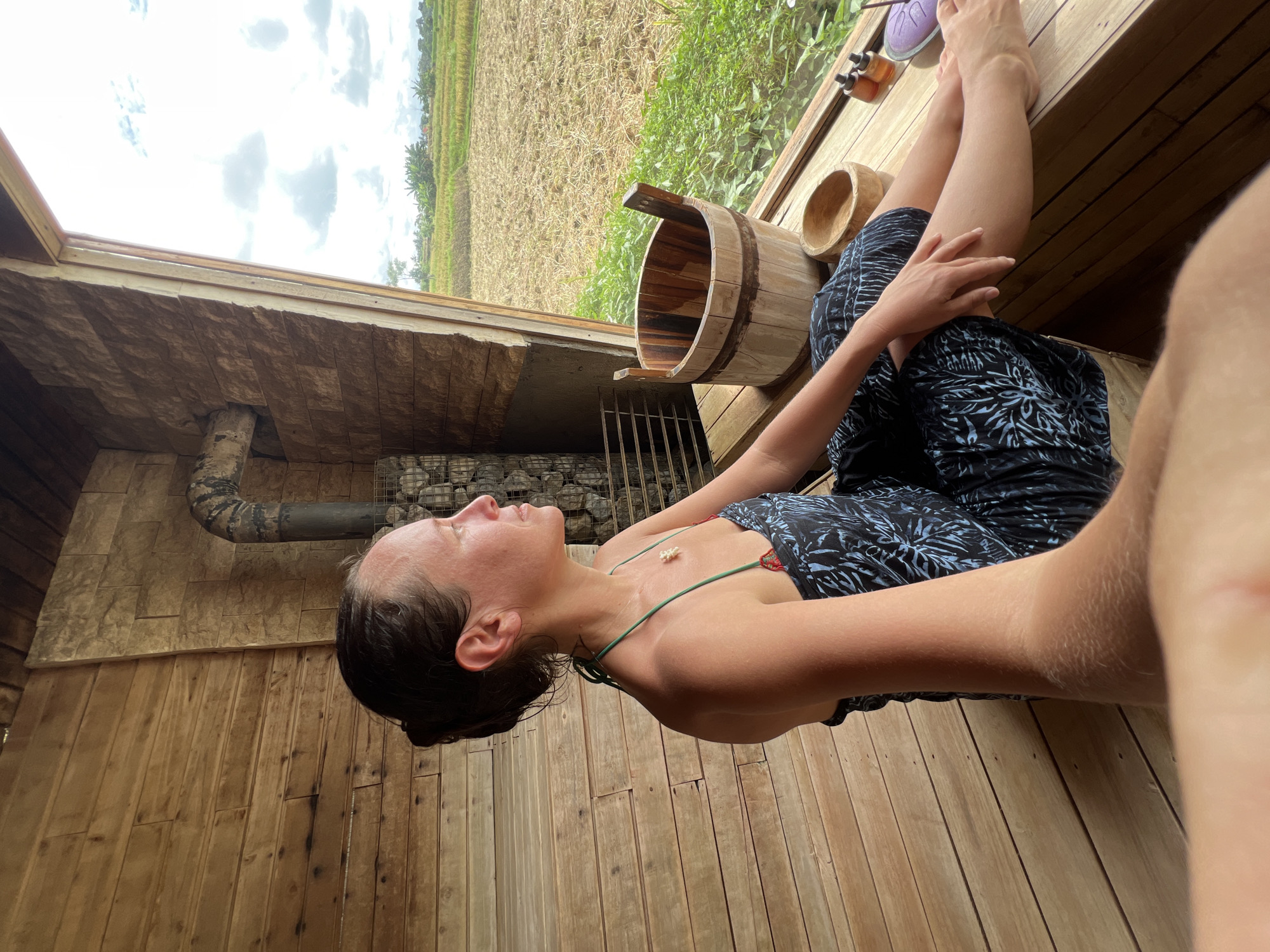 A woman relaxes in a sauna located in the Ubud District, Indonesia, as evidenced by the nearby rice paddies visible through an opening. She is reclining with her legs crossed, wearing a patterned dress with thin straps. A wooden bucket and bowl sit nearby. The sauna has wooden walls and a stone-covered wall with a pipe running across it, presumably for heating.
