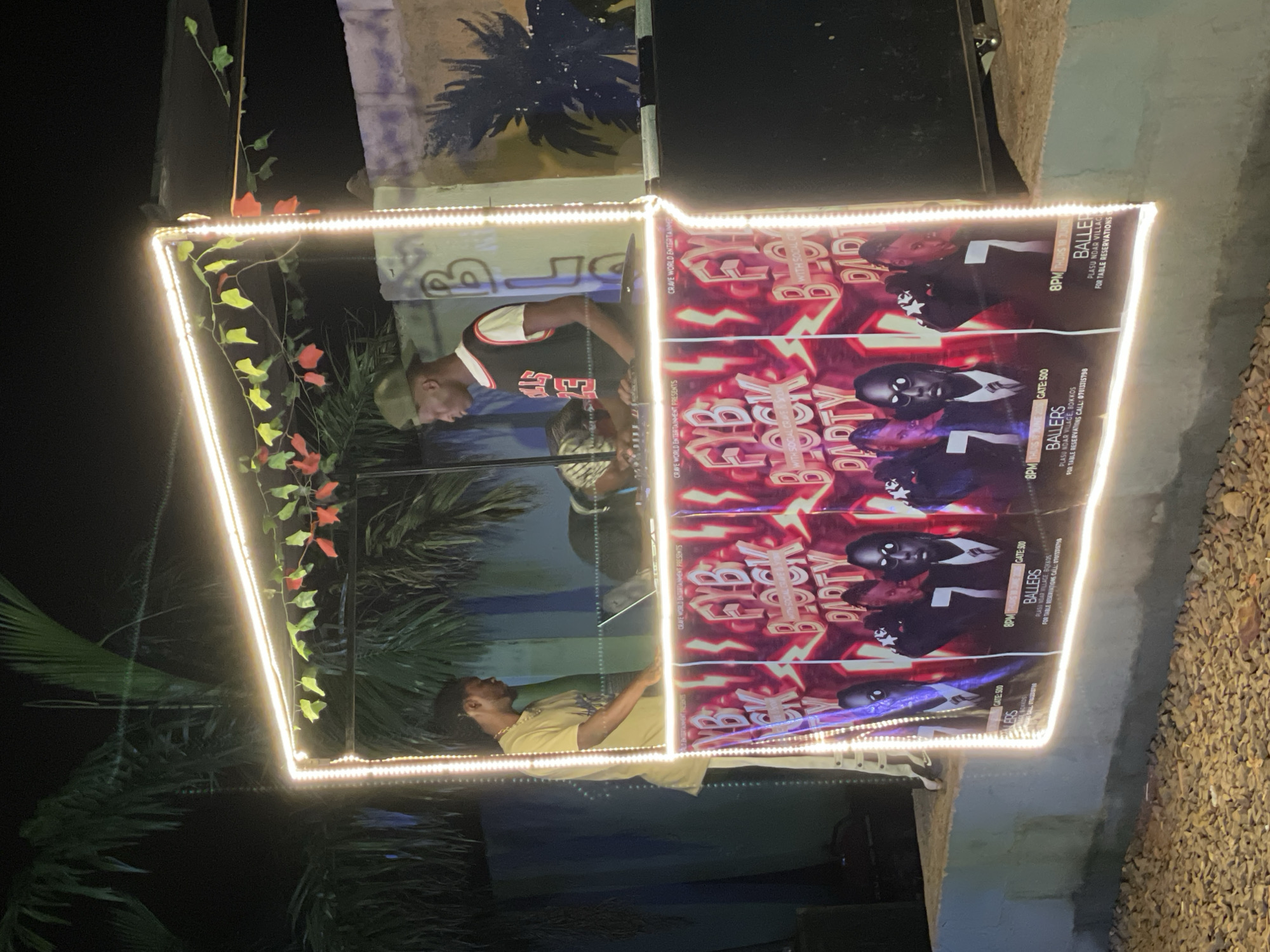 An outdoor nighttime scene in Durumi, Nigeria, features a makeshift DJ booth illuminated by a string of white LED lights. Two young Black men are present. One man, wearing a light yellow t-shirt and cream-colored pants, stands to the left, facing a laptop on the DJ console. The second man, wearing a dark sleeveless basketball jersey over a white t-shirt and a green cap, is positioned behind the console, appearing to operate the equipment. In front of the DJ booth, a banner advertising an event titled "FYB BLOCK PARTY" is displayed, featuring images of several men. The background includes palm trees and a wall with graffiti. The ground in the foreground is covered in gravel.