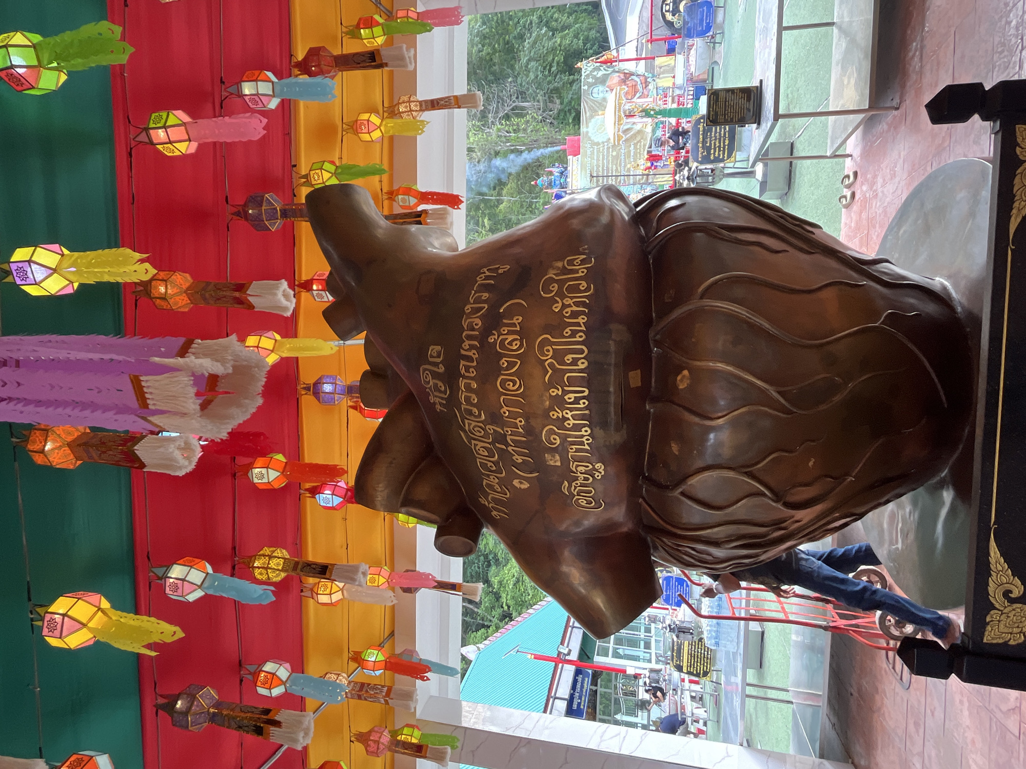 Here is a description of the image:
The image shows a close-up of a bronze statue with gold inscriptions in Thai script. The statue is positioned against a backdrop of colorful hanging lanterns in shades of green, red, yellow, pink, and blue. The inscriptions appear to be part of a dedication or description of the statue. In the background, outside a covered area, there is a glimpse of green vegetation, a white building, and other structures that suggest a public space. People can be seen in the background, but they are not the main focus of the image. The location is the city of Chao Phraya Surasak, Thailand.