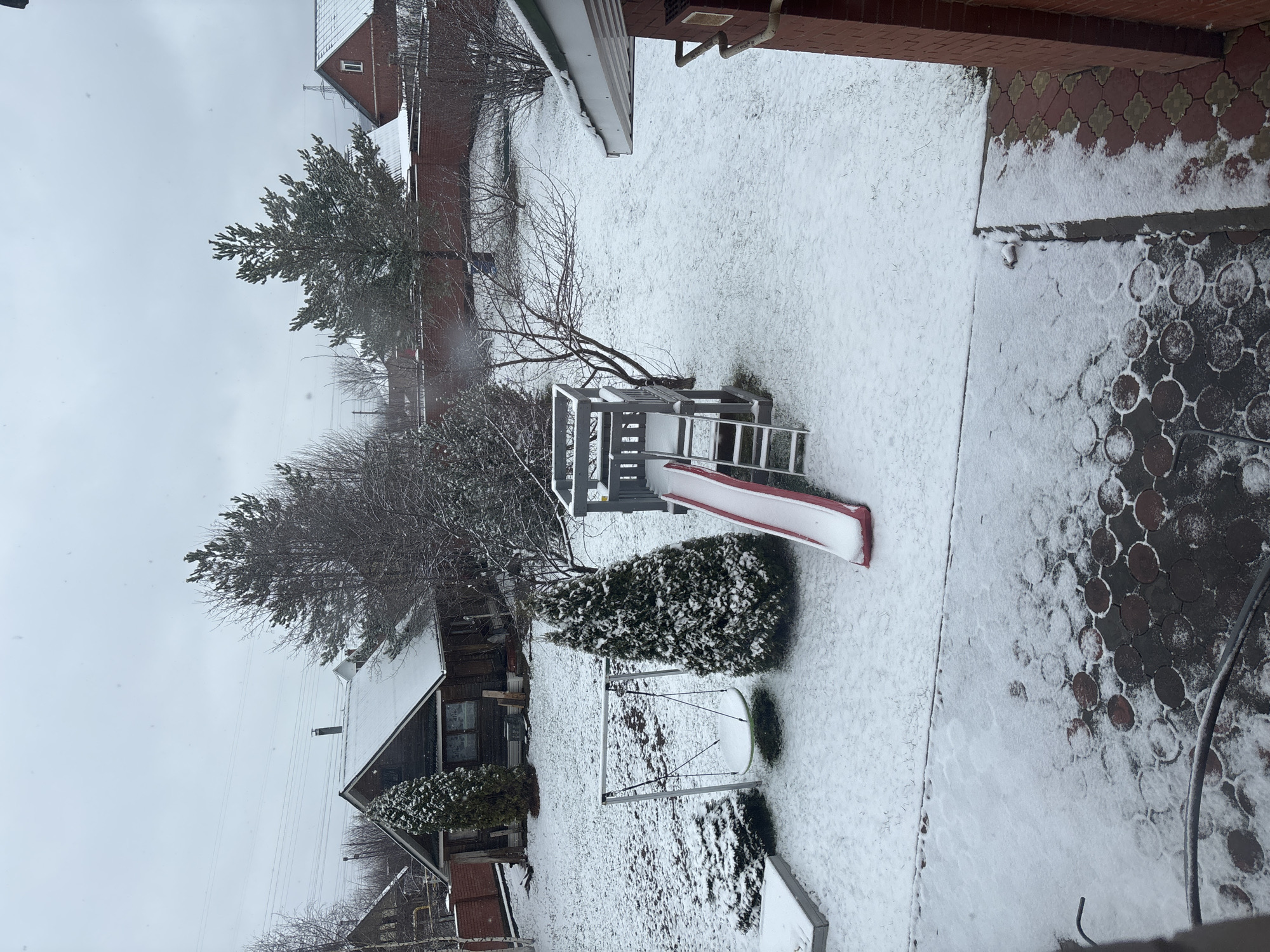 The image shows a snow-covered backyard in Ферма, Russia. A children's playset with a red slide is partially visible. A swing set and a few evergreen bushes are also present. A house is visible in the background, and a brick-paved area borders the yard. Snow is falling. No people are visible.