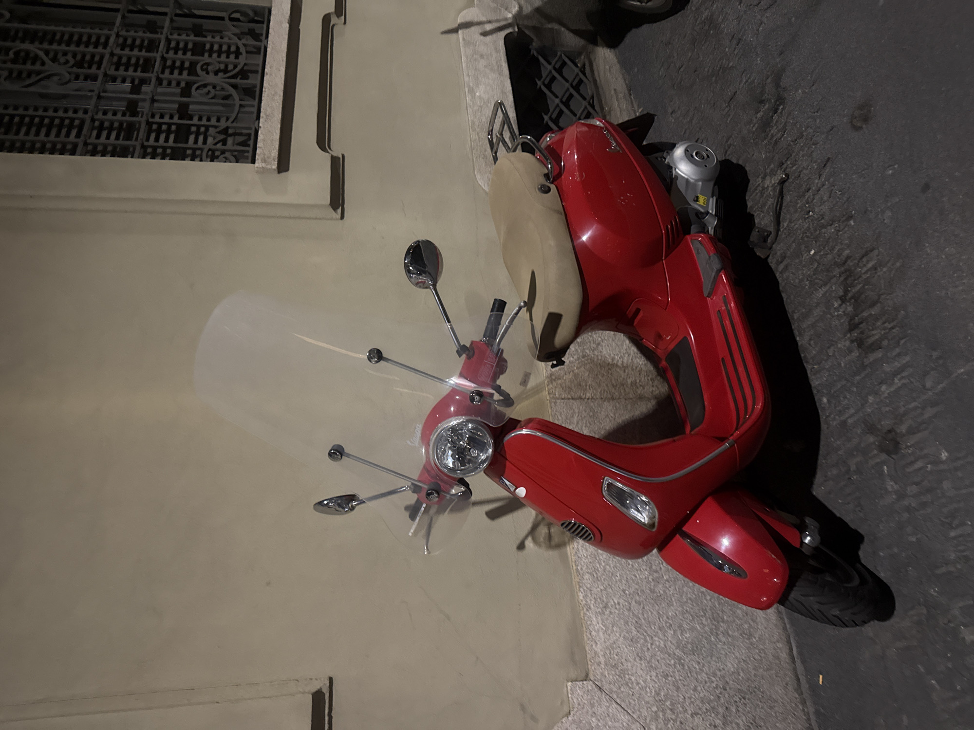 The image displays a vibrant red Vespa-style scooter parked vertically on a street or sidewalk, likely at night or during dusk, in Milan, Italy. The scooter is the primary subject, featuring a glossy red body, a contrasting beige seat, and a tall, clear windshield. Its handlebars are chrome, adorned with multiple chrome mirrors that reflect the ambient light, and a prominent round headlight is visible at the front.
The scooter is positioned next to a light-colored building facade, which appears to be made of stucco or stone. Part of a window with an intricate, dark wrought iron grate is visible towards the top left, hinting at classic European architecture. The ground beneath the scooter is a mix of textured light-colored pavement and darker asphalt, indicating a city street and sidewalk. Deep shadows are cast by the scooter onto the street, suggesting illumination from an artificial light source like a streetlight, which highlights the scooter's bright color against the darker surroundings. No individuals are present, and no discernible text is visible on the scooter or the building. The scene captures a characteristic urban snapshot, common in Italian cities.