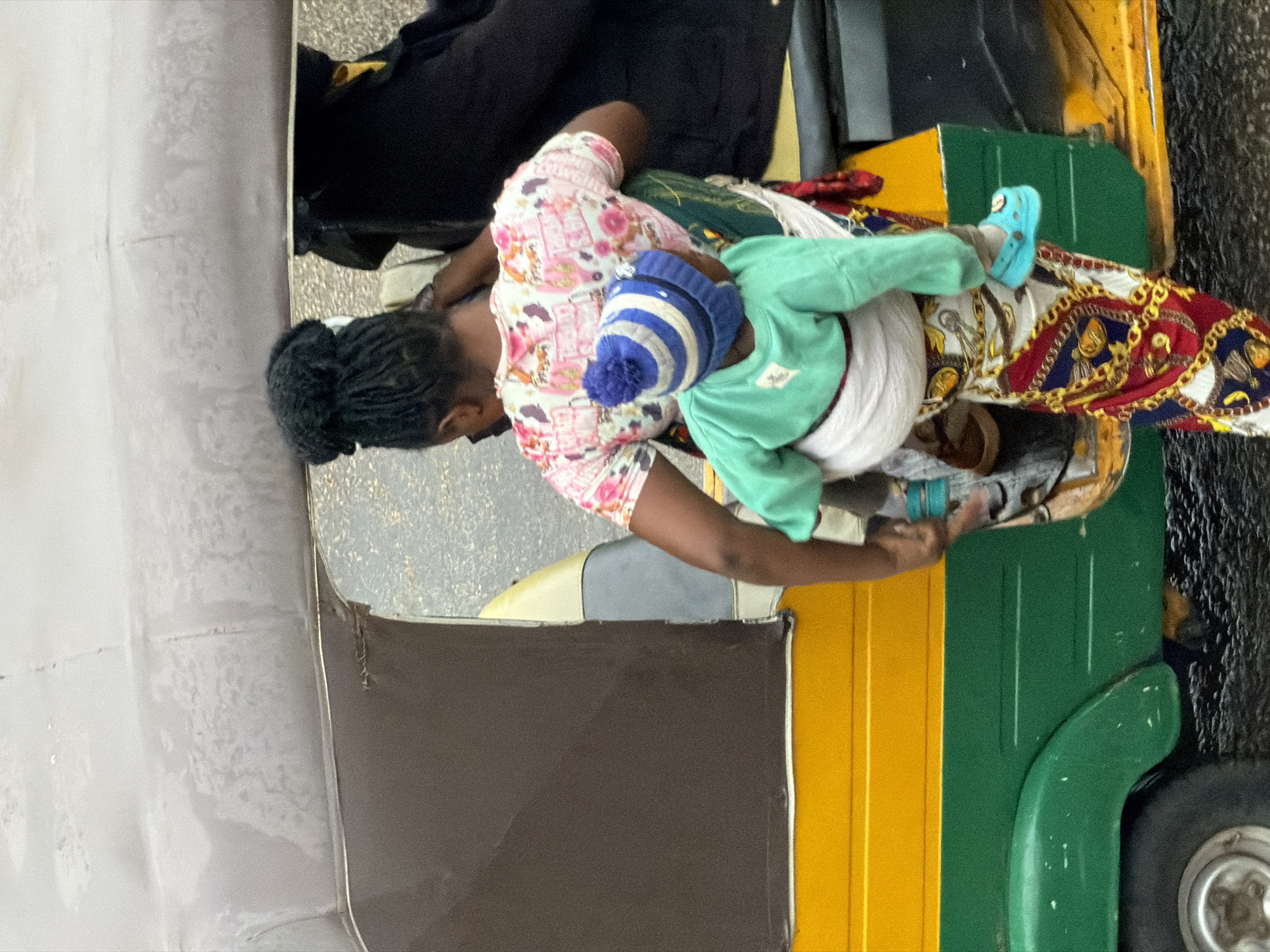 A woman in a pink floral-print shirt and a colorful patterned skirt, carrying a child on her back, stands next to a yellow and green auto-rickshaw. The child wears a blue and white striped hat and a mint green sweater. The woman's hair is styled in dark, braided locs. The auto-rickshaw has a brown fabric covering on the top. The ground appears to be wet and dark, suggesting it may have recently rained. The image is taken outdoors in what appears to be a street setting, possibly in the city of Bukuru, Nigeria.