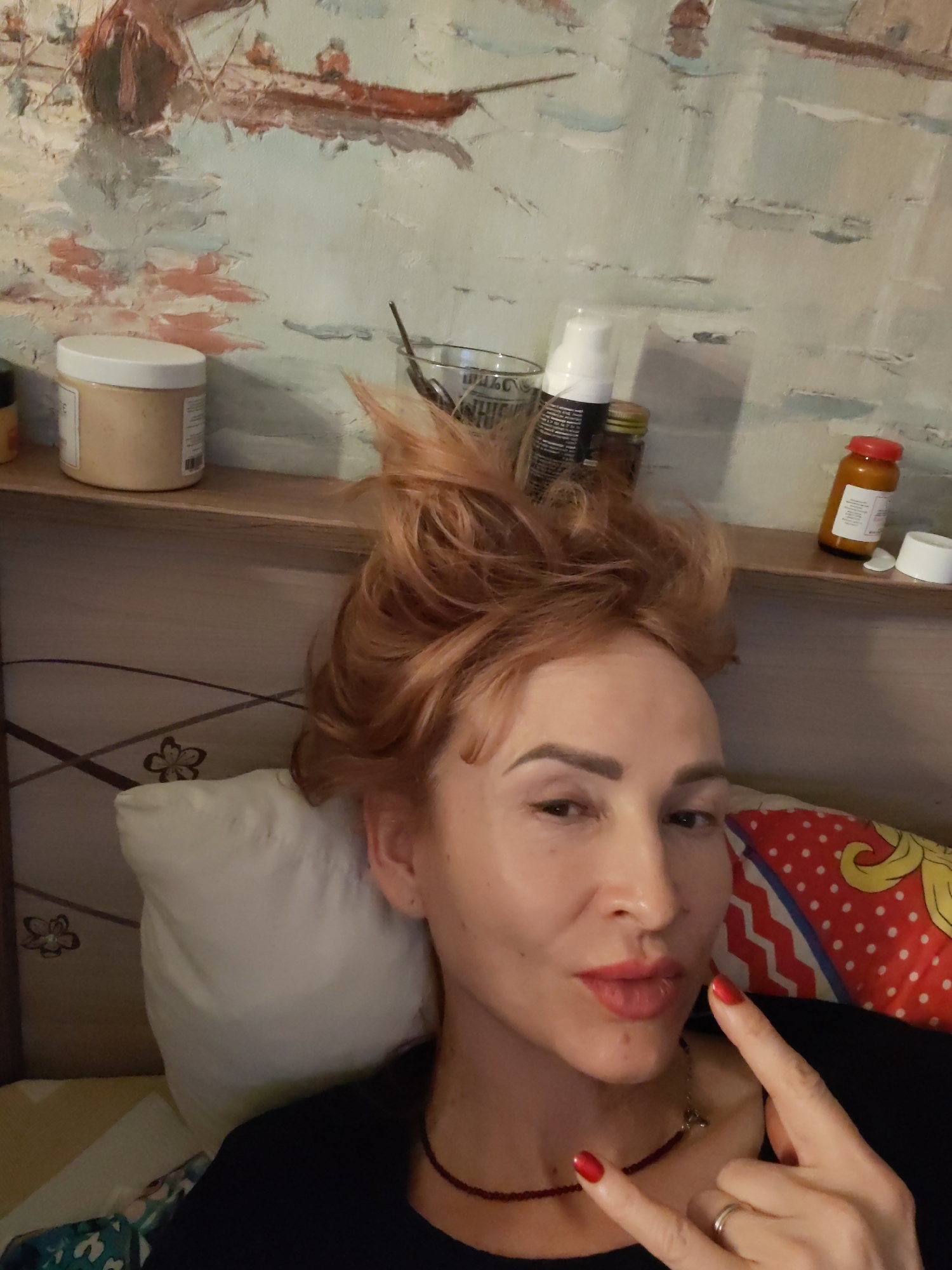 The image shows a person with reddish-orange hair lying on a bed. The person is wearing a black top and has a beaded necklace. The person is making a gesture with their fingers.
Behind the person is a headboard with a design and a shelf. On the shelf are several bottles, jars, and containers. There is a painting on the wall above the shelf depicting boats on water.
There is no information in the image to indicate a specific location in the city of Chaikovsky, Russia.