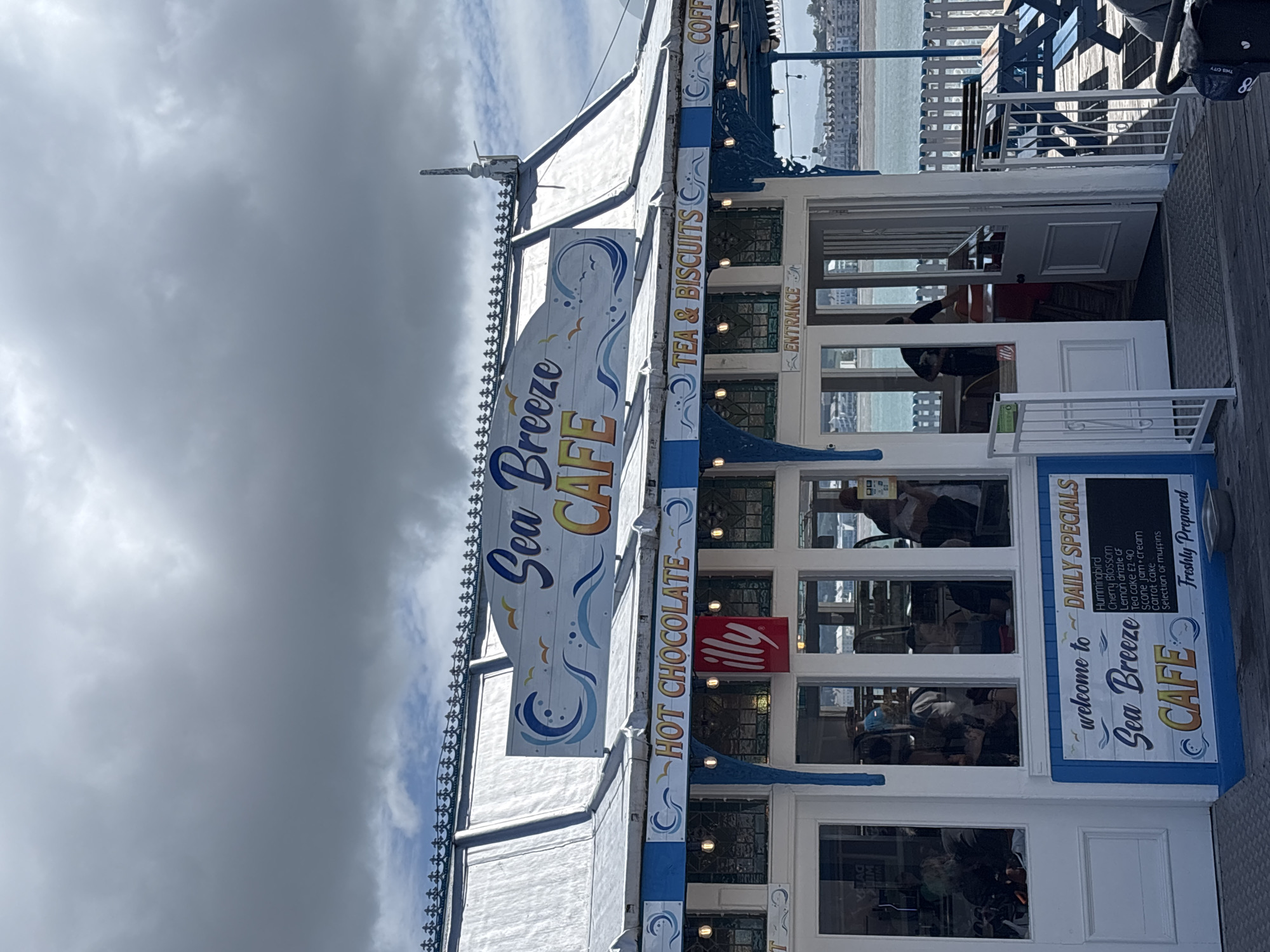 The image depicts the exterior of the "Sea Breeze CAFE" in Llandudno, United Kingdom. The cafe is a white building with blue accents and is adorned with signs advertising hot chocolate, tea, biscuits, and coffee. One sign prominently displays "Sea Breeze CAFE" along with wave motifs. Another sign lists "Daily Specials," including Hummingbird and Cherry Blossom options. Through the windows, people can be seen seated inside. The cafe appears to be situated on a pier or boardwalk, with the sea and the Llandudno coastline visible in the background. The sky is overcast with heavy grey clouds.
