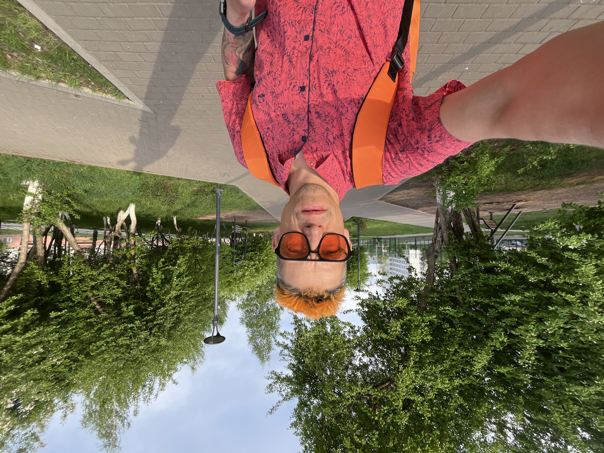 A selfie photograph shows a man outdoors in Chaikovsky, Russia. He is wearing orange-tinted sunglasses, a patterned red shirt, and orange suspenders. He stands on a paved path surrounded by trees and grass. A building is visible in the background. Streetlights are also present.