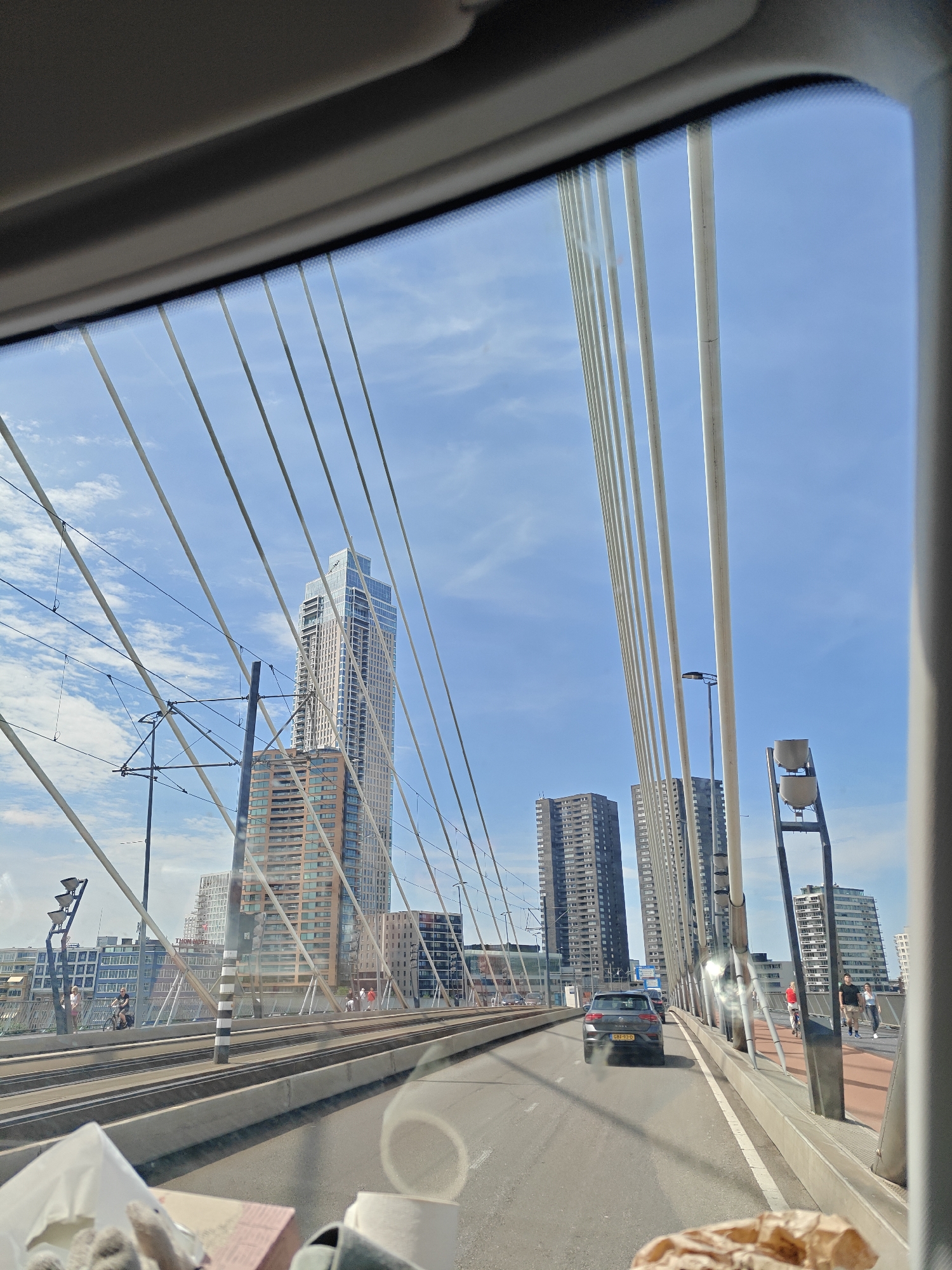This media file captures a view from inside a vehicle, likely a car, looking through the windshield. The scene depicts a cable-stayed bridge with multiple tall, slender white cables stretching upwards against a bright blue sky with scattered white clouds. The perspective is from the road deck of the bridge, looking forward.
Several modern high-rise buildings are visible in the background, showcasing a city skyline. One prominent building is a tall, slender skyscraper with a light-colored facade and numerous windows. To its right, two darker, blockier skyscrapers stand side-by-side. The bridge itself appears to have multiple lanes of traffic and also includes tram tracks running parallel to the road.
Several people are visible on the pedestrian walkway of the bridge. Some are walking, while one person is riding a bicycle. A dark-colored hatchback car is driving in the lane ahead of the viewer's vehicle.
The time of day appears to be daytime, with bright sunlight casting some reflections and glare on the windshield and surfaces. The weather seems pleasant, with a clear blue sky indicating good visibility. The overall atmosphere is one of urban transit and daily life within a modern cityscape.
The license plate of the car in front is partially visible and reads "GBF-97-D". A black and white striped pole is also visible on the bridge. In the foreground, on the dashboard or console of the vehicle, there are some crumpled paper objects, possibly napkins or bags, and a white roll that could be toilet paper or paper towels.