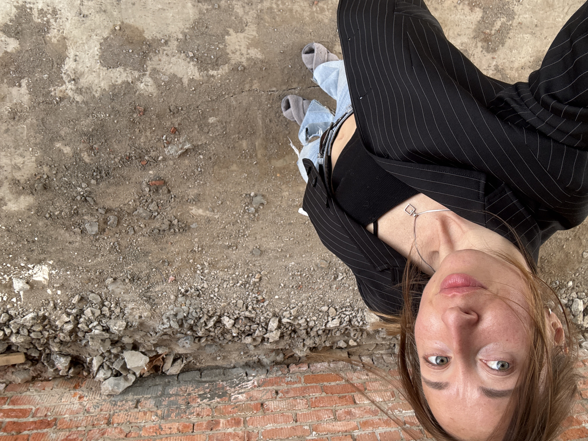 A female person, viewed from a high angle, stands on a rough, broken surface of exposed brick and concrete rubble. She wears a black pinstripe blazer, a black top, and ripped denim jeans. The setting appears to be a derelict or under-construction area in Chaikovsky, Russia, judging by the unmaintained environment. The woman wears grey socks with sandals. A necklace is visible around her neck. The picture composition makes the background appear to be the floor.