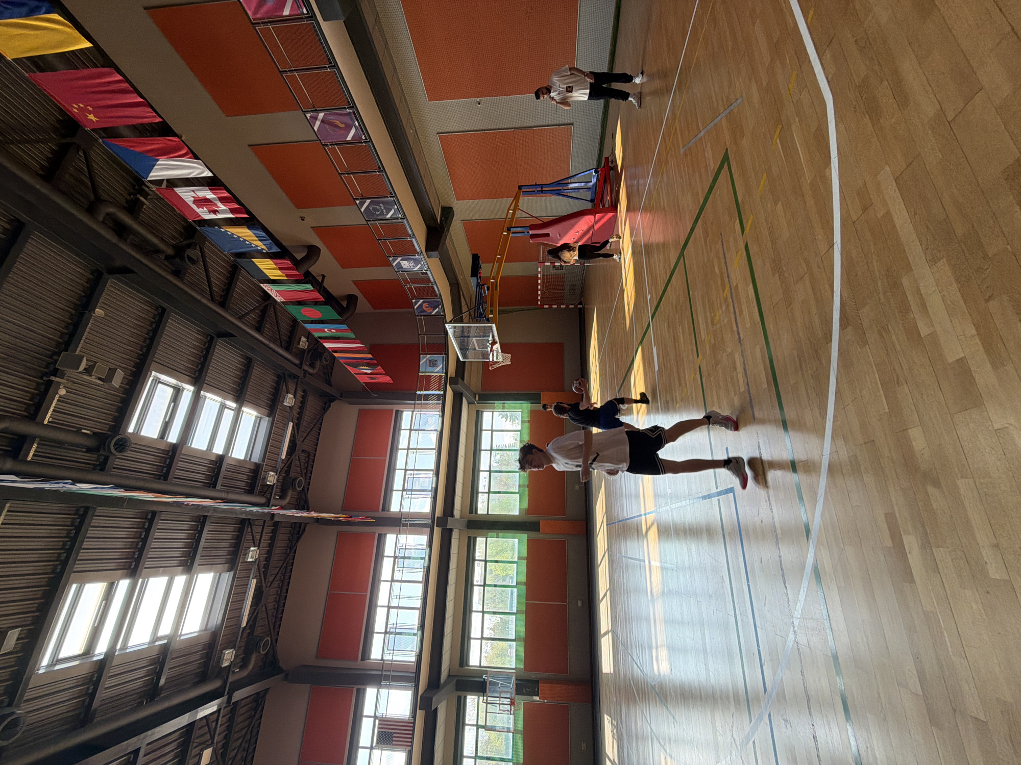 The image shows the interior of a gymnasium in Blagoevgrad, Bulgaria. The room is brightly lit by several rows of windows along one wall, with a dark ceiling structure above. A series of flags representing various countries are suspended from the ceiling. There are several people in the gymnasium. One person is standing near a basketball hoop on the far side of the room, and another is located close by. The gymnasium floor is made of wood. A person wearing a white shirt and black shorts is stretched out on the floor.