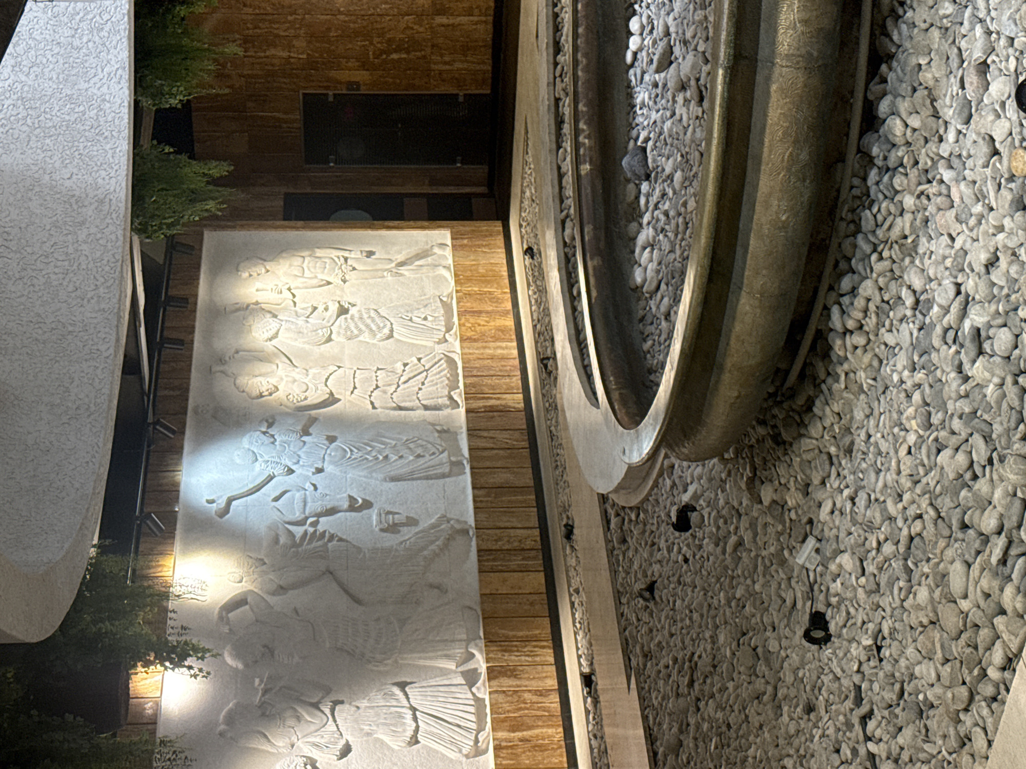 The image shows a circular fountain with a stone basin, filled with small, grey pebbles. The fountain is surrounded by more grey pebbles on the ground. In the background, there is a textured white wall with a bas-relief carving of figures, possibly depicting a historical or mythological scene. Above the carving, there is a curved ceiling with hanging greenery. To the right of the wall, there is a wooden panel and two dark doorways. The lighting is dim, with spotlights illuminating the bas-relief. The overall scene suggests an indoor or covered outdoor public space, possibly a hotel lobby, museum, or park. The location is identified as Yalta, Ukraine, suggesting this is a feature within the city.