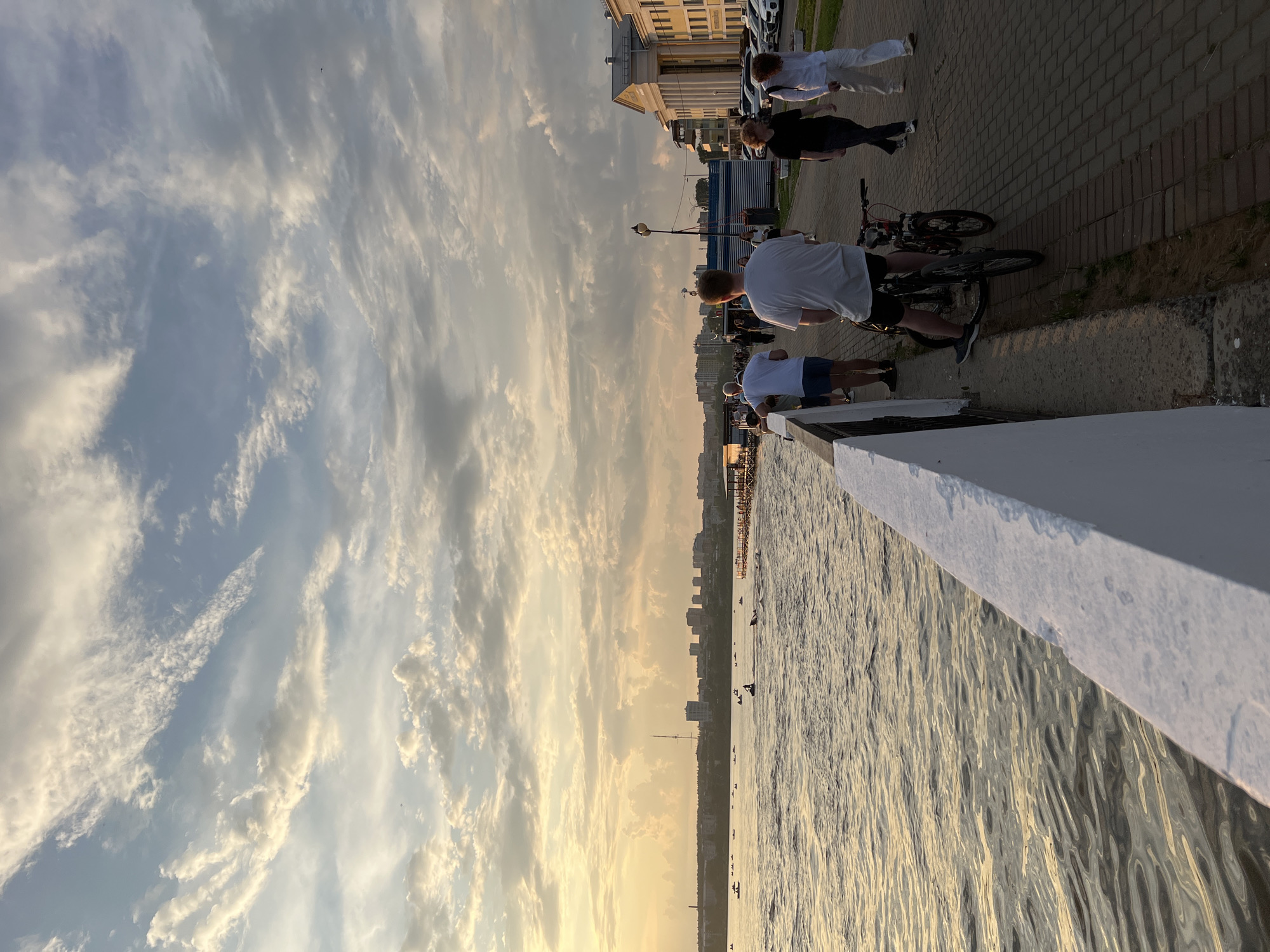 The image depicts a waterfront promenade in Izhevsk, Russia, during a sunset. The sky is filled with dramatic clouds, illuminated by the setting sun, creating a warm, golden glow. On the left side of the image, the water reflects the sky, with ripples indicating a gentle breeze. Several small boats and what appear to be individuals on personal watercraft are visible on the water in the distance.
The promenade itself is paved with brickwork and lined with a low white railing. Several people are walking along the promenade. In the foreground, a man is standing next to a bicycle, with another bicycle in front of him. Further along the promenade, a couple is walking away from the camera, and to their right, a woman and another person are walking towards the camera.
On the right side of the promenade, a large building with classical columns and a yellow facade is visible. Cars are parked in front of this building. In the background, a city skyline with various buildings, including what appears to be a tower, is silhouetted against the illuminated sky. The overall atmosphere is one of a pleasant evening spent by the water.