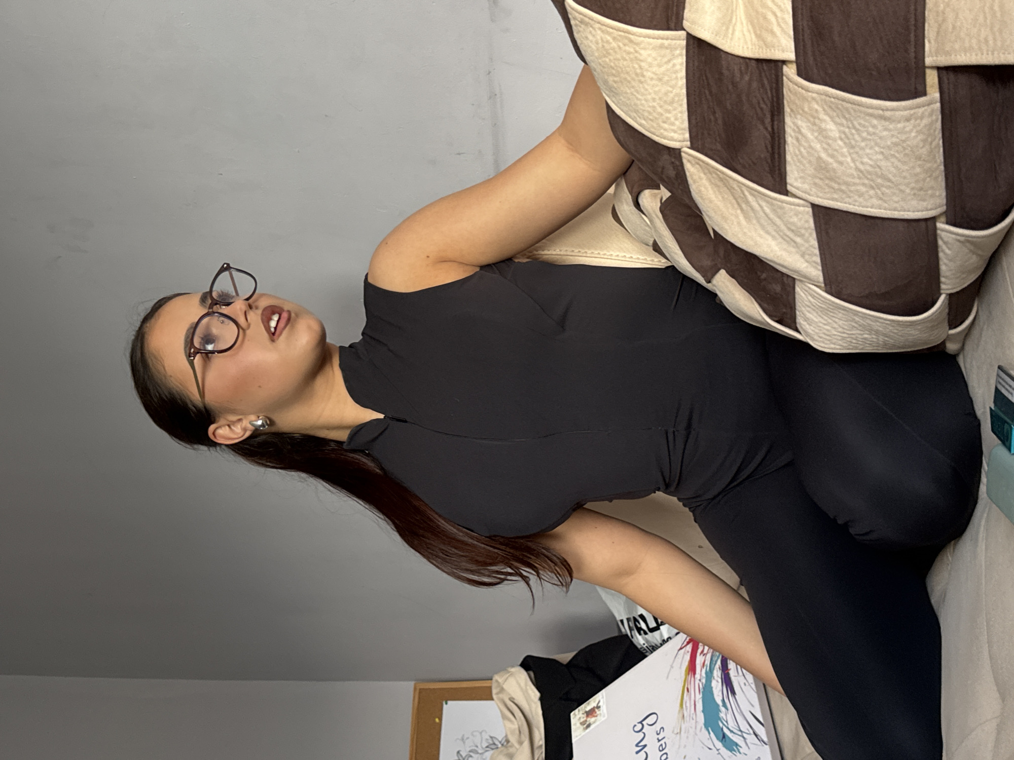 A woman with long dark hair and glasses is leaning against a large brown and beige checkered pillow, which is positioned on a light-colored surface that may be a couch or bed. She is wearing a black top and black leggings. The background includes a beige corkboard with papers attached, other miscellaneous items on a light surface, and a light gray wall. The location is Unknown, Unknown.