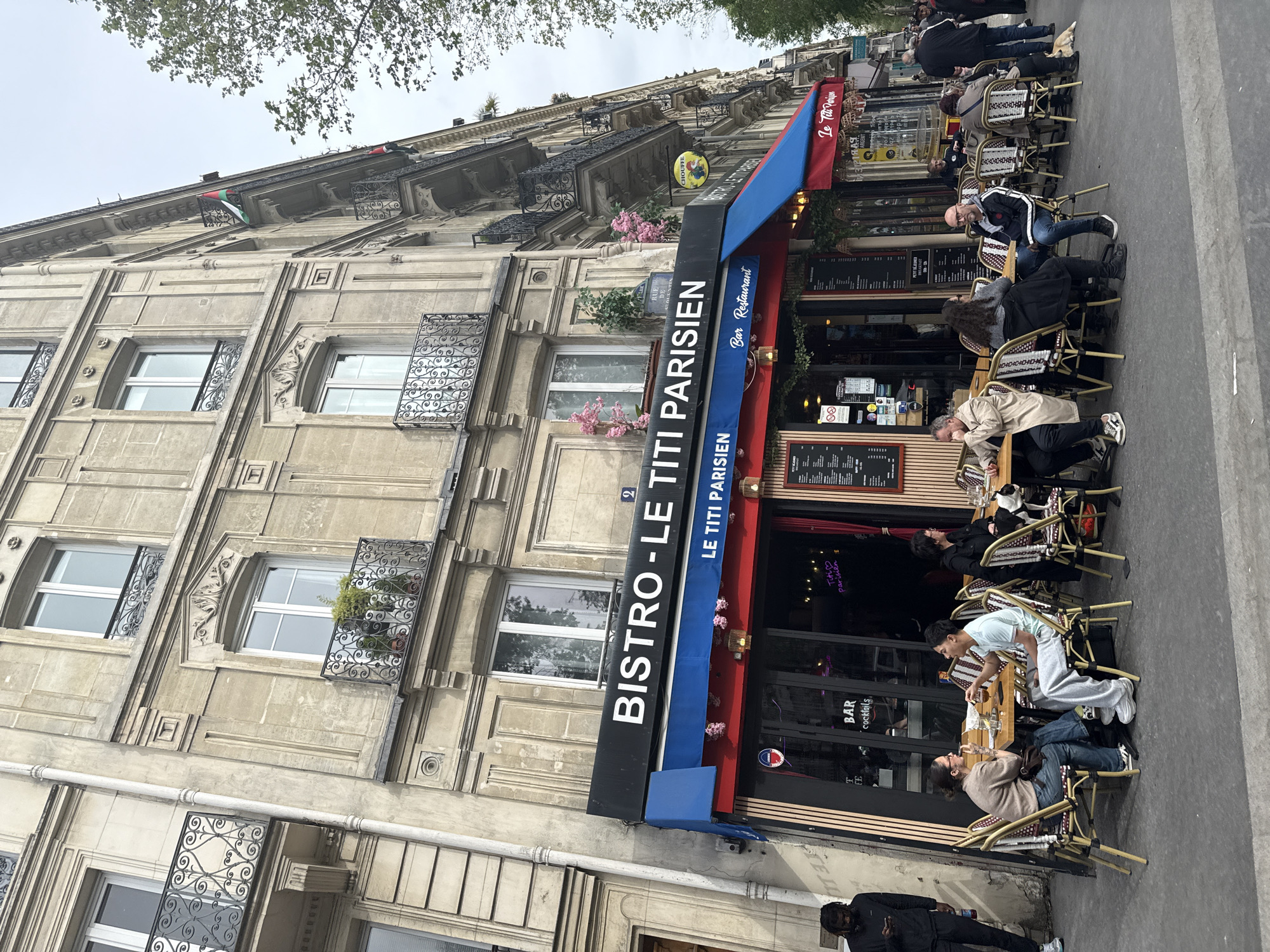 The image depicts the exterior of the "BISTRO-LE TITI PARISIEN," a restaurant in Paris, France. The restaurant has a blue and red awning and outdoor seating occupied by patrons. The building is of classic Parisian architecture, with several windows featuring ornate ironwork. People are seated at the tables, some are talking, and others are looking at their phones. A person is walking on the sidewalk in front of the restaurant. The environment appears to be a typical Parisian street with trees visible in the background. A flag hangs on the building's roof.