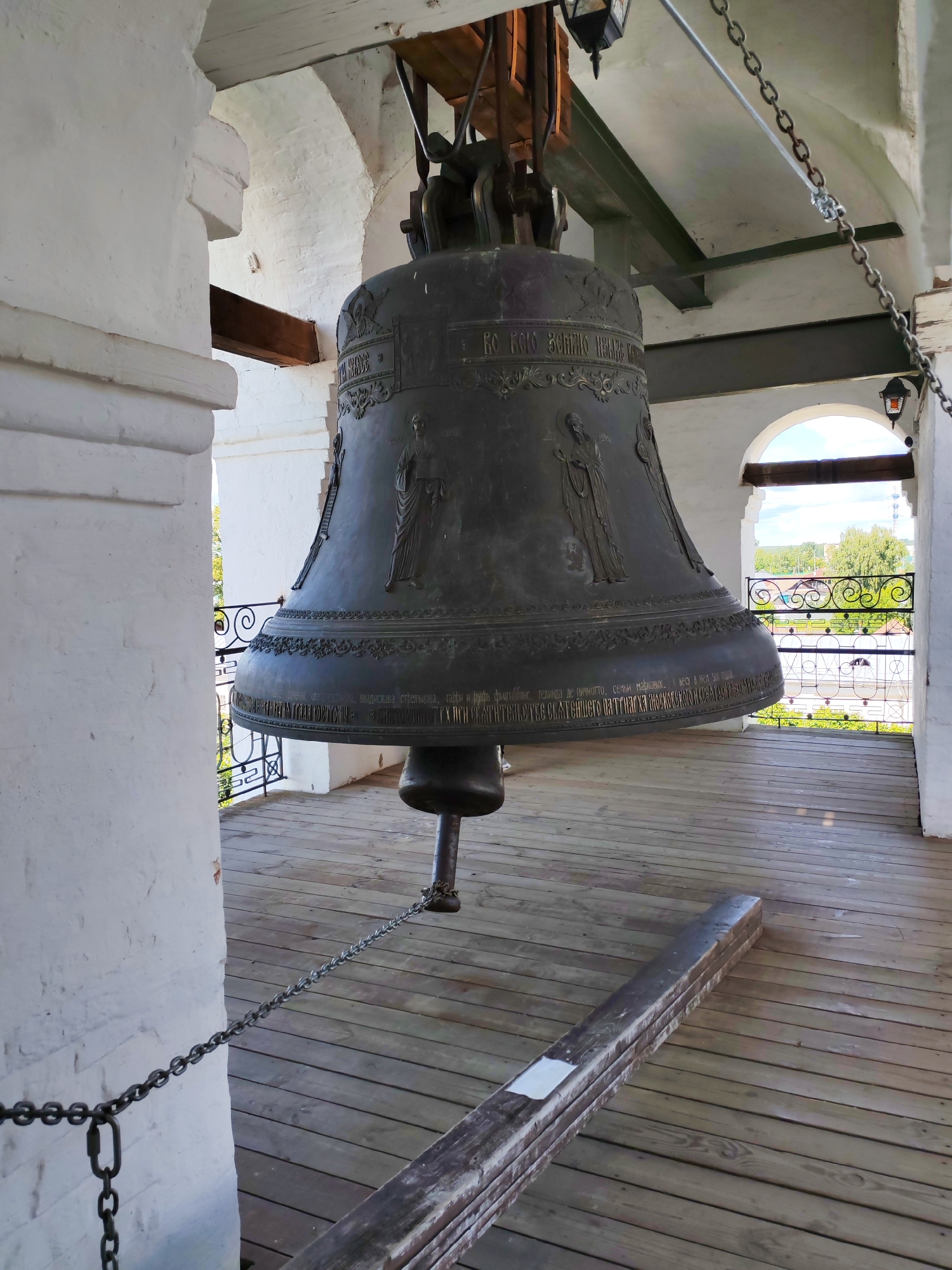 This media file shows a large, ornate bronze bell hanging inside a bell tower, likely in Kostroma, Russia, given the location context. The bell is decorated with bas-relief images of figures, likely religious in nature, and inscriptions. It is suspended from a wooden and metal support structure.
The bell tower features white brick walls and a wooden floor. A metal chain is visible, attached to the bell and extending down to the floor, likely for striking or controlling the bell. Through an arched opening in the background, a scenic view of greenery, trees, and rooftops can be seen, suggesting it is daytime with fair weather. The overall scene conveys a sense of historical and religious significance, with the imposing bell as the central subject. The visible text on the bell appears to be in Cyrillic script.