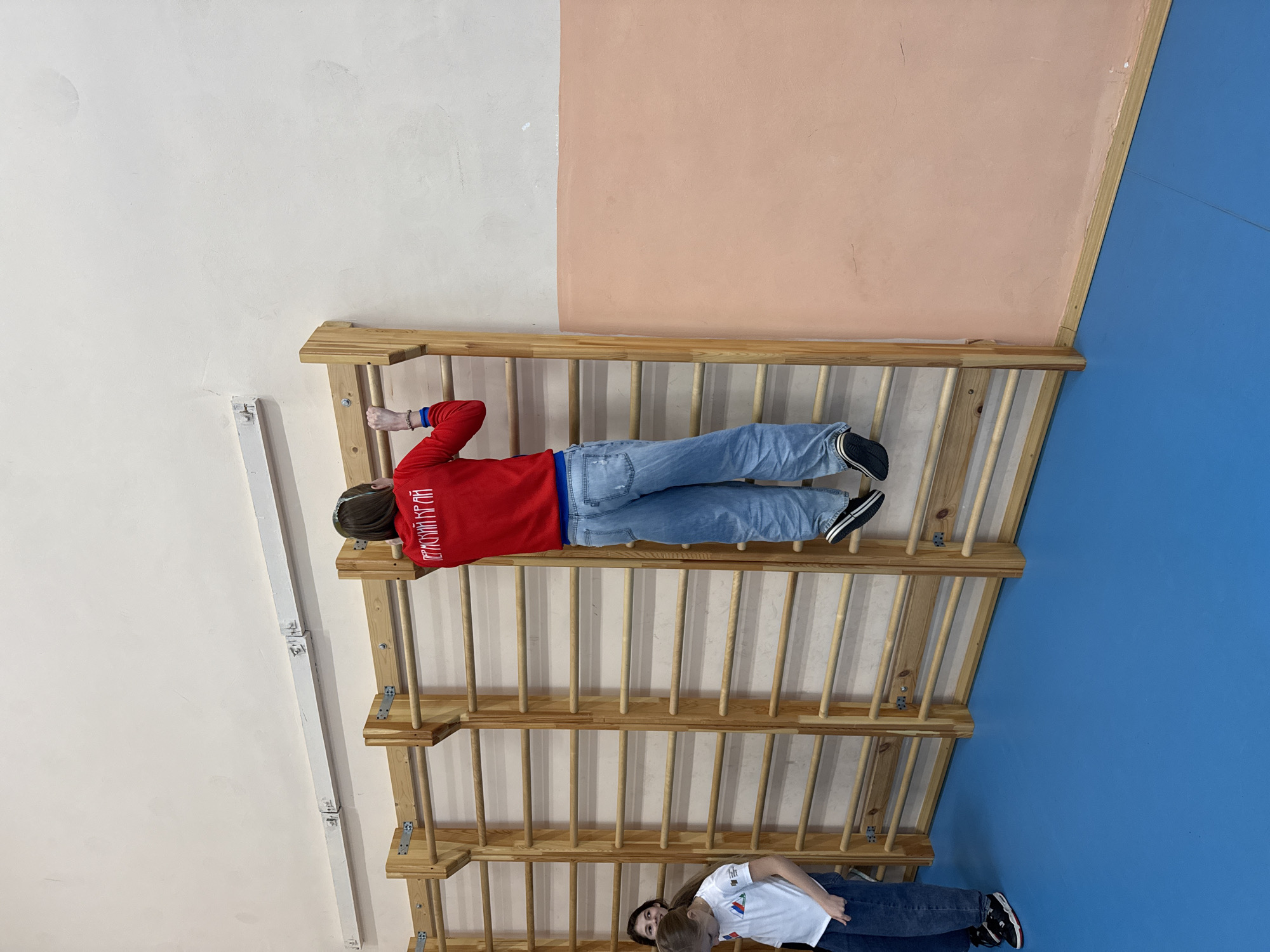 A man wearing a red sweater and jeans is lying horizontally on a wooden ladder-like structure. Another person is visible at the bottom of the ladder structure, and the background consists of three wall panels colored white, peach, and blue. The image was taken at the Museum of Illusions in Usolye, Russia.