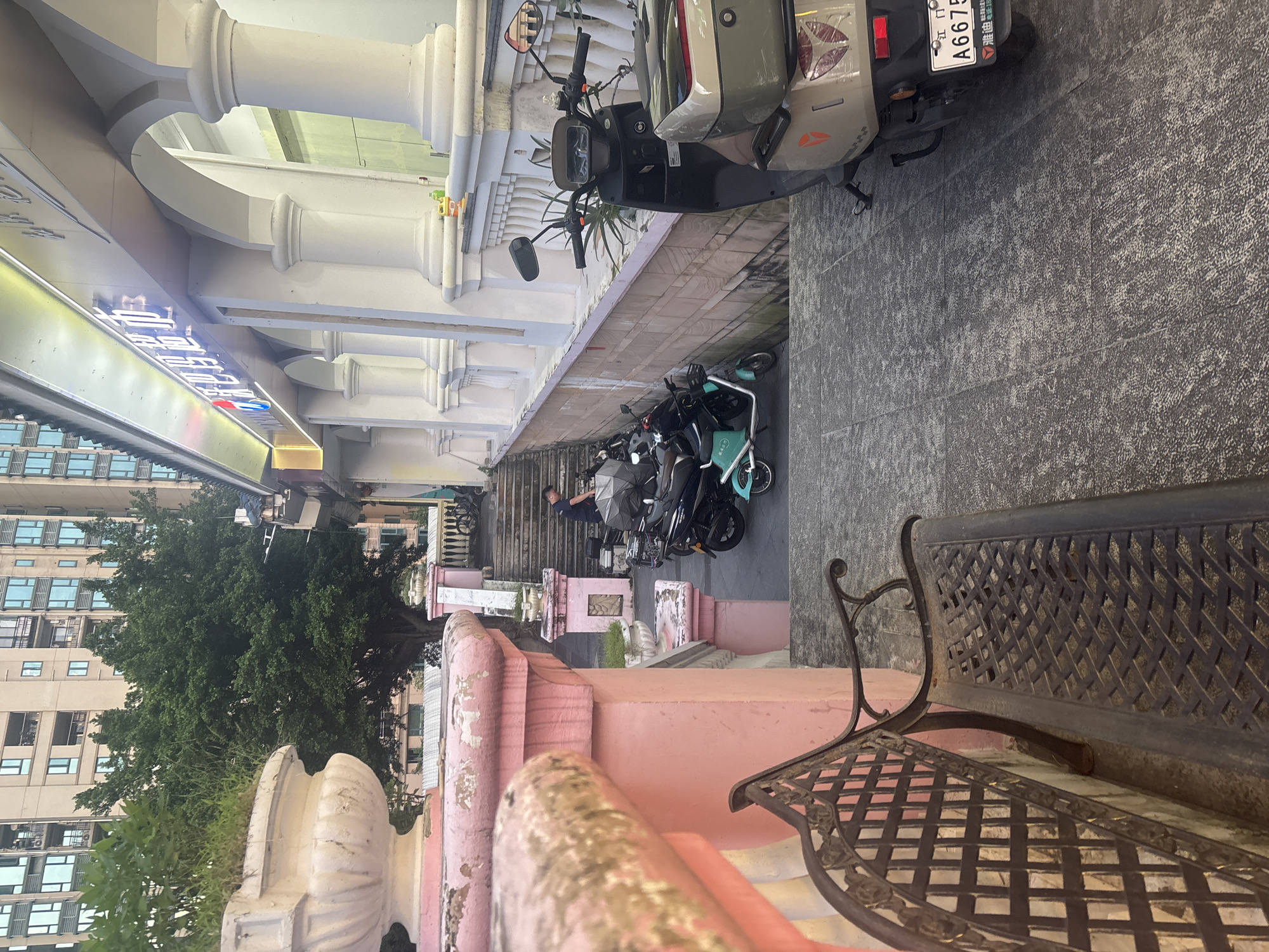 In Nanhai District, China, a stairway leads down from a building with an arched facade and decorative pillars. The building's signage is illuminated. Parked on the stairs and pavement are several electric scooters and motorcycles. A person in a dark shirt stands near some of the scooters. In the foreground, a weathered metal bench faces the stairway. A large planter with greenery sits on a pink, ornate structure to the left, overlooking the scene. Tall residential buildings are visible in the background, along with trees.