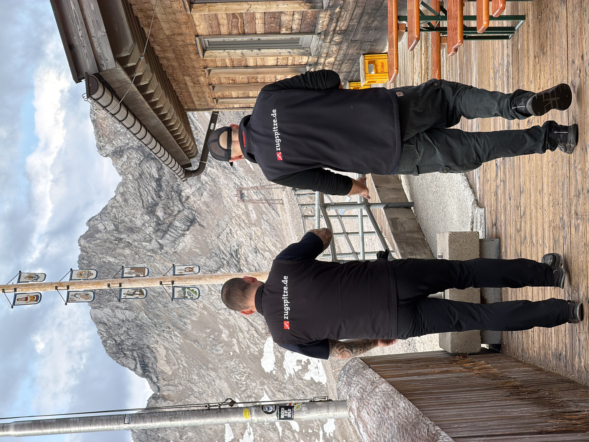 The image shows two men walking away from the camera on a wooden boardwalk. Both men are wearing black vests with a red and white logo that reads "zugspitze.de". The man on the left has tattoos on his arms. The man on the right is wearing a black baseball cap. Behind them, a rustic wooden building with windows and a slanted roof is visible. A tall wooden pole with several small flags hangs on the left side of the image, and in the background, a large, rugged mountain range with patches of snow rises against a cloudy sky. The scene appears to be at a high-altitude location, likely the Zugspitze, Germany's highest peak, near Garmisch-Partenkirchen. Wooden tables and benches are set up to the right of the boardwalk.
