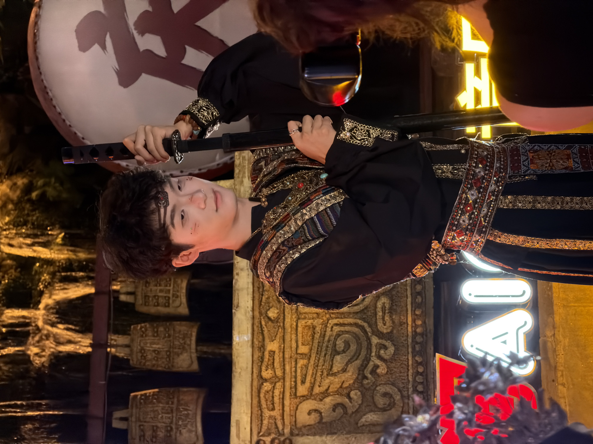 In Yuzhong District, China, a young East Asian male stands wearing a black, traditional-style outfit adorned with intricate gold embroidery and colorful beaded detailing on the collar, sleeves, and a wide belt. He holds a black sword vertically in his right hand. The background features a row of antique-looking bells hanging above a golden, ornate structure with carved designs. To the right, a large drum with red calligraphy is visible. In the lower portion of the image, a bright yellow neon sign with Chinese characters, partially obscured, illuminates the scene. The overall atmosphere suggests a cultural performance or themed event.
