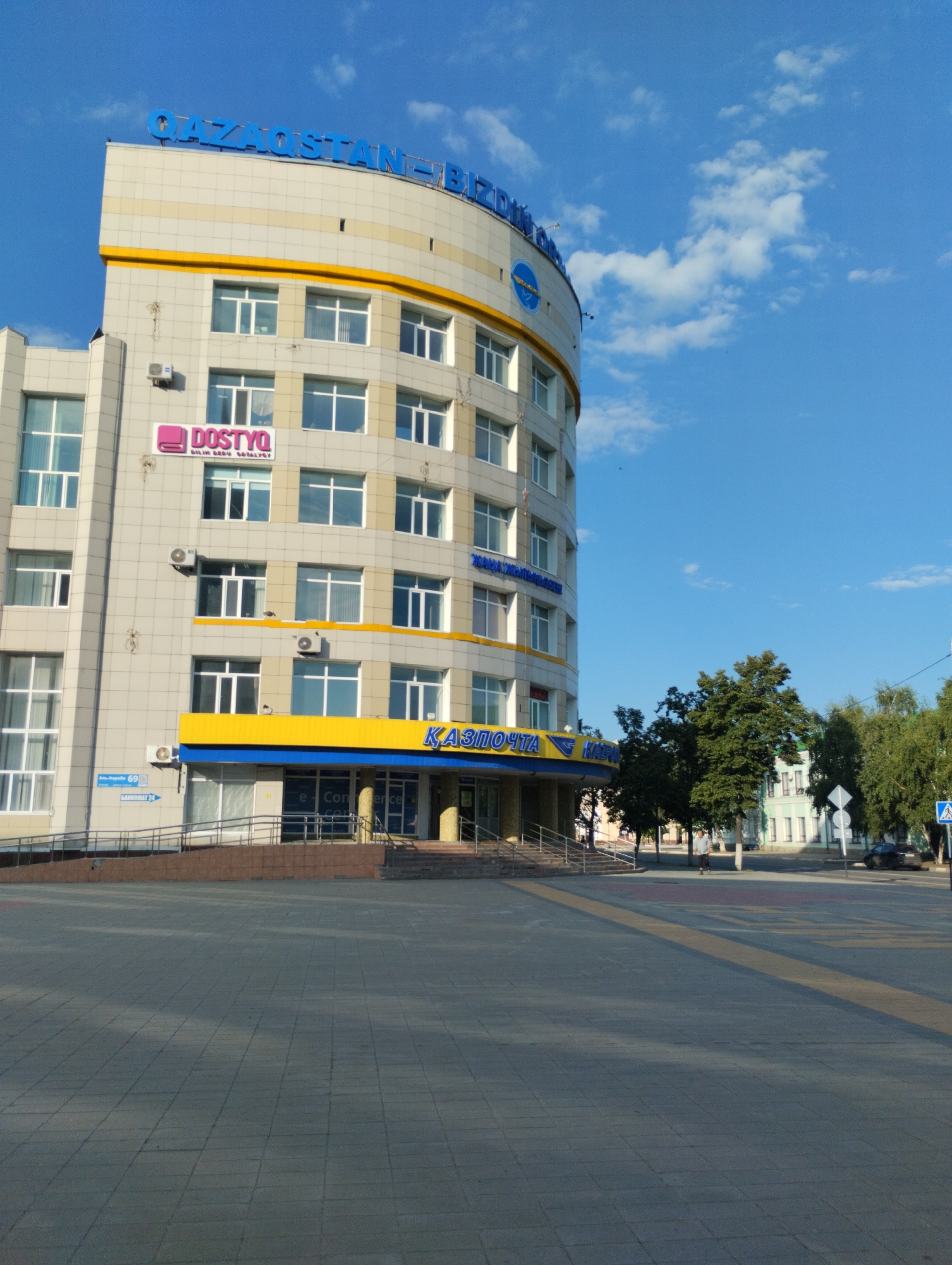 The image shows the Qazaqstan-Biznes-Center building in Kostanay, Kazakhstan. It is a multi-story building with a curved facade, predominantly light-colored with yellow and blue accents. The building has numerous windows and air conditioning units. On the lower floors, there is signage indicating "DOSTYQ" and "KAZPOSTA." The building is situated on a paved plaza with trees and other buildings visible in the background. The sky is blue with scattered clouds. A pedestrian crossing and a car are visible on the street in front of the building. There are no people prominently featured in the image, though a person is walking in the background.