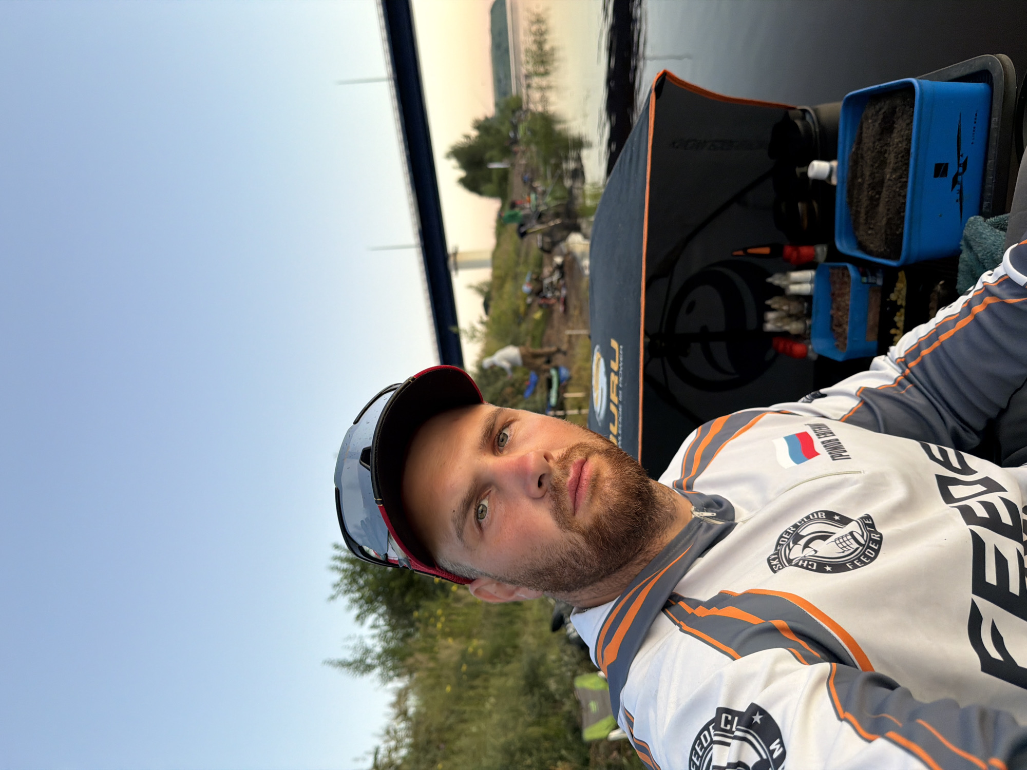 The image shows a man wearing a fishing vest and a baseball cap, with sunglasses perched on the cap. He is looking at the camera. In the background, there is a body of water, a bridge, and trees. The man appears to be at a fishing spot. The location is identified as Tarasovo, Russia. The scene is set in what appears to be a natural environment, possibly a riverbank or lakeside, with the sky suggesting it is either sunrise or sunset. Several other people are visible in the background, also seemingly engaged in fishing activities, with fishing gear and tents set up along the shore. There are containers of fishing bait and other supplies near the man.