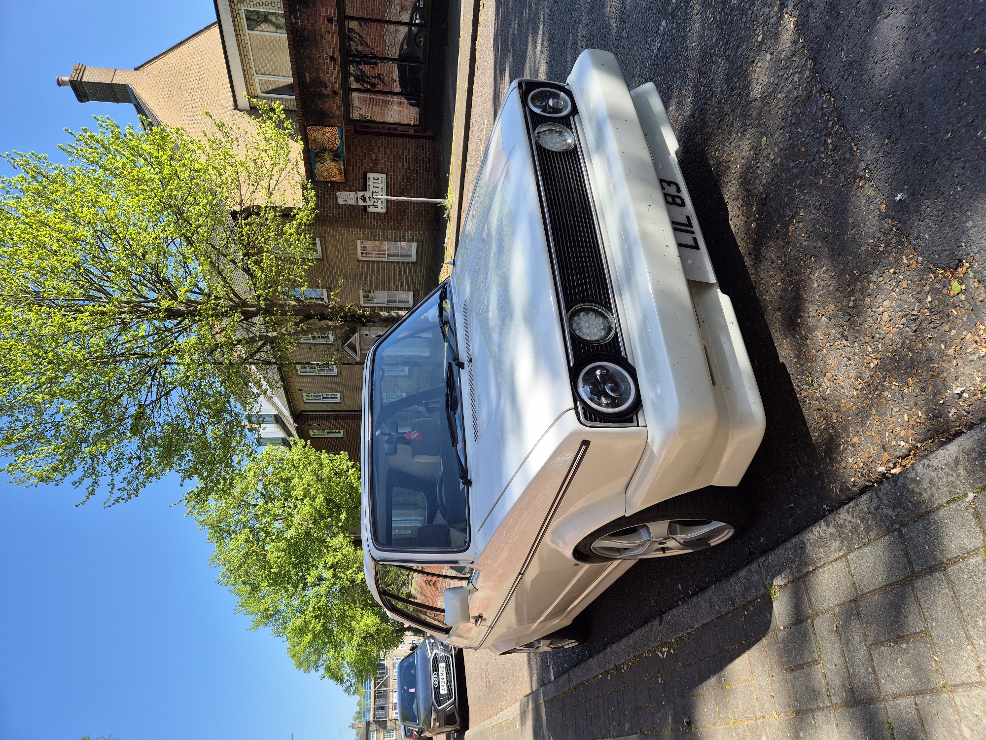 This image, taken in West Suffolk, United Kingdom, captures a white classic car, likely a modified Volkswagen Golf Mk1, parked on a paved street. The photograph is taken at an oblique angle, with the car diagonally traversing the frame from the lower left to the upper right.
The car is the central subject, featuring a white body, a black front grille, and modern circular LED headlights. A thin, subtle brown stripe runs along the side of the vehicle. Its custom front license plate clearly displays "L1L 83". Behind the white car, a darker vehicle, possibly a sedan, is partially visible, with its license plate showing "AV24 MHZ".
To the left of the parked cars, a large, mature tree with vibrant, fresh green leaves fills a significant portion of the scene, indicative of spring or early summer. Beyond the tree, a traditional British building constructed of reddish-brown brick with numerous windows and a prominent chimney stands under a clear, bright blue sky. A white sign with visible text "METRIC" is affixed to the building.
The street is asphalt, with a paved sidewalk of grey square slabs visible to the right of the car. Bright sunlight illuminates the scene, casting distinct shadows from the car and trees onto the street and sidewalk, suggesting a clear, sunny day, likely mid-morning to late afternoon. There are no people visible, and the overall impression is one of a calm, everyday street scene.