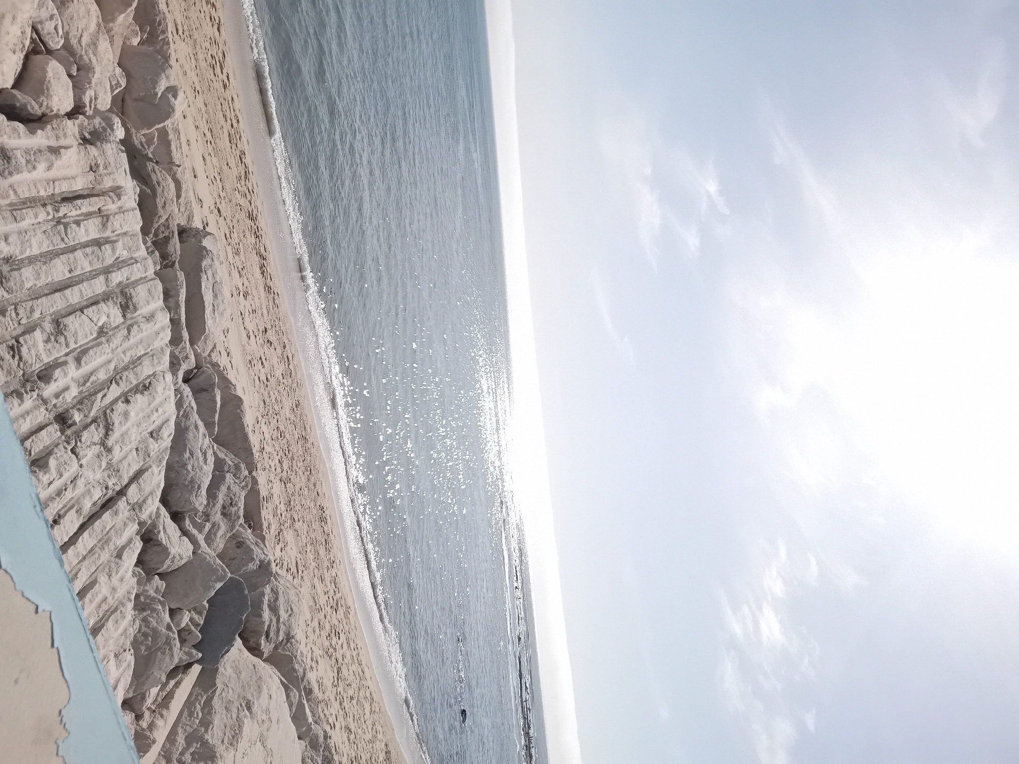 A seaside scene displays a beach with light tan sand bordered on one side by an expanse of water glistening with sunlight and on the other by a rocky area featuring cut stone. The water transitions into a bright sky filled with scattered clouds. An element painted pale blue is visible along the lower left edge of the image. The image is rotated, tilting the horizon line diagonally. The location is not identified.