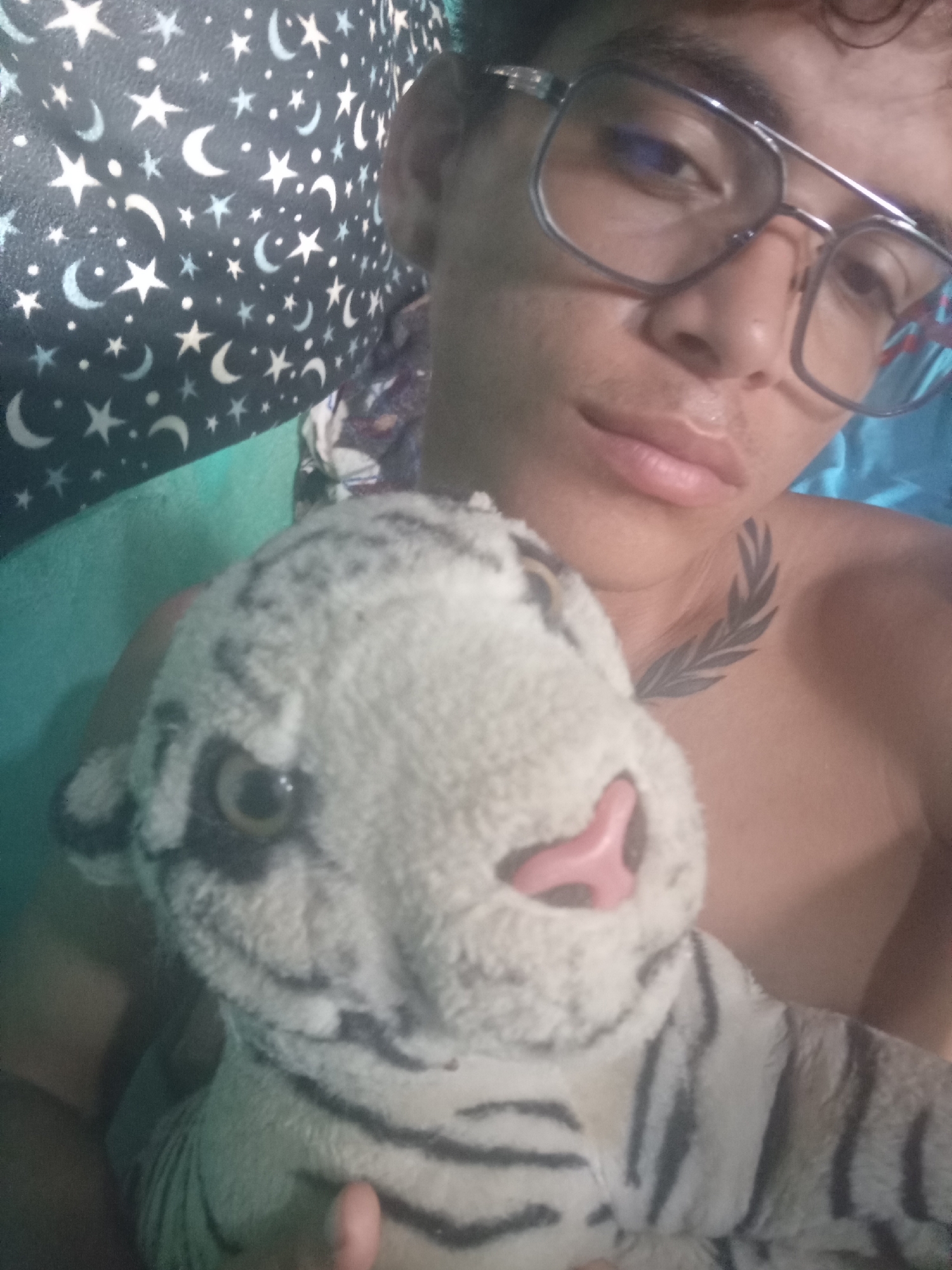 This is a medium shot, eye-level, selfie-style photo taken indoors. The subject, a young person with curly dark hair, is wearing large, square-rimmed glasses and is shirtless. They are holding a stuffed toy tiger with a light-colored body and black stripes. The toy tiger's face is in the foreground, with its pink nose and yellow-brown eye visible. The background includes a turquoise wall and a dark fabric with a pattern of white stars and crescent moons. A tattoo of a leafy branch is visible on the subject's neck, below their jawline. The lighting appears to be artificial and soft, casting gentle shadows.