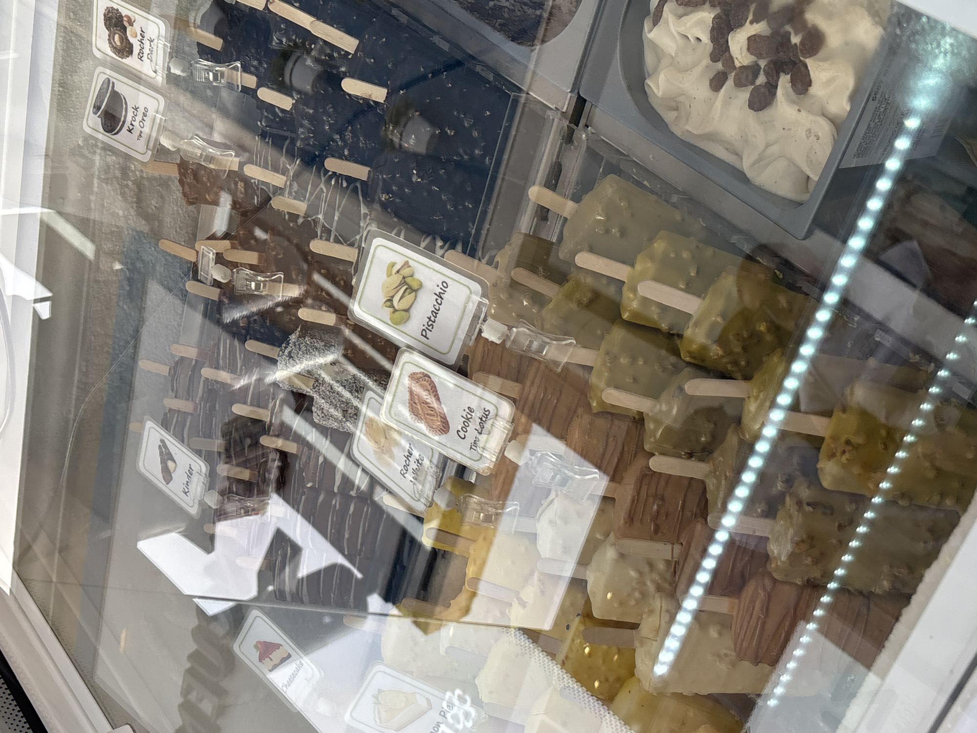 The image shows a display case filled with various ice cream bars and tubs of gelato. The ice cream bars are individually wrapped or coated and arranged in rows, with small printed labels indicating their flavors. Some of the visible flavors include "Kinder," "Cheesecake," "Krock," "Rocher Dark," "Rocher White," "Cookie," and "Pistacchio." The tubs of gelato are also visible, with one containing a creamy white gelato with raisins or nuts. The display case appears to be in an ice cream shop. The reflections on the glass suggest an outdoor or well-lit indoor environment. There are no people visible in the image. The lighting is bright, illuminating the products clearly. The overall impression is of a well-stocked ice cream parlor. Based on the provided context, this ice cream shop is located in the city of Orihuela, Spain.