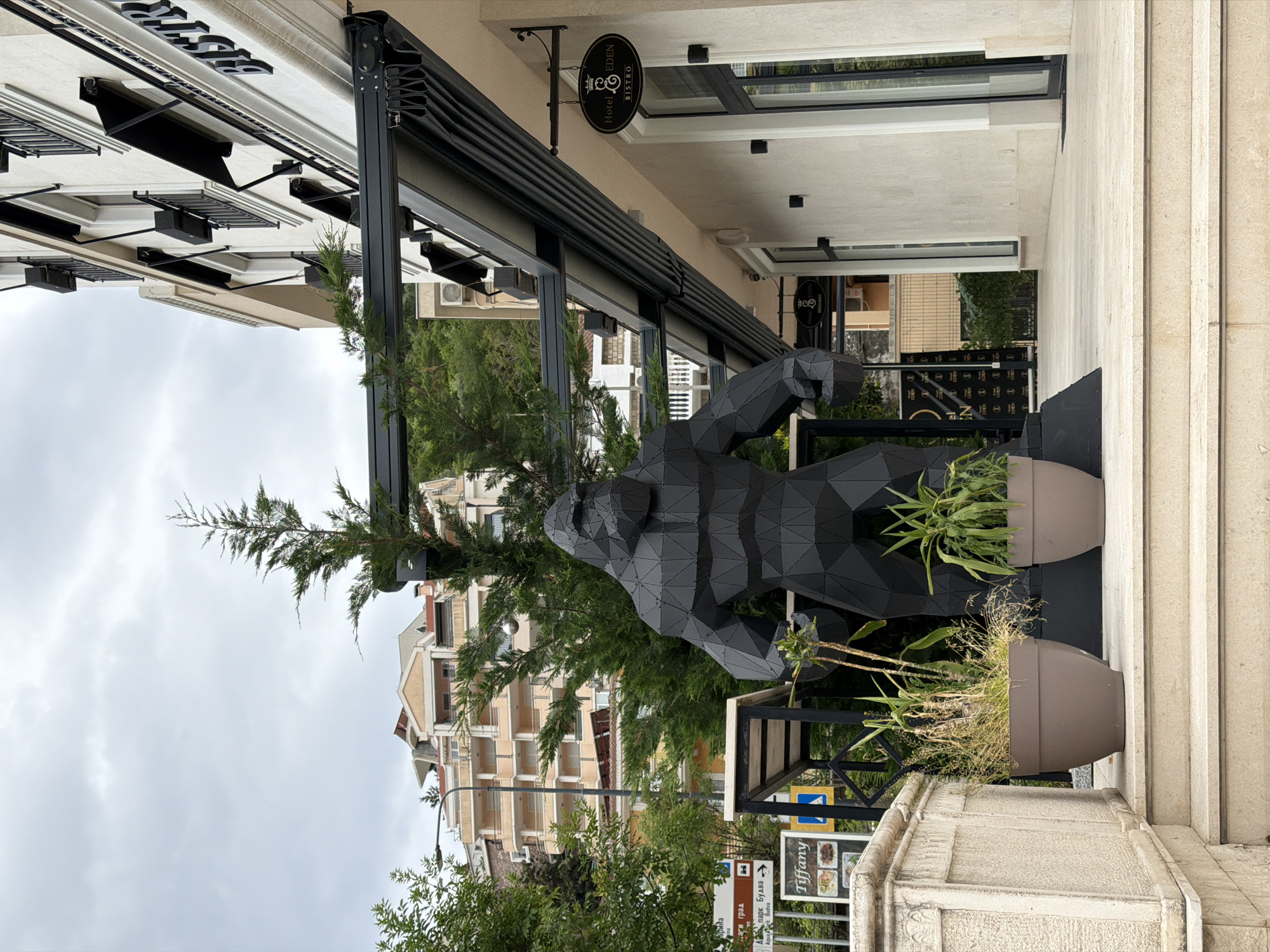 The image shows a low-angle view of a sculpture of a gorilla, apparently made of black geometric plates, standing in front of the Hotel Eden Bistro in Budva, Montenegro. The sculpture is placed between two large, light-brown planters with greenery. Behind the sculpture, a building with light-colored facades and balconies rises, partially obscured by the branches of a coniferous tree. To the left, the sky is overcast. A sign is visible at the bottom, displaying "Tiffany" with an arrow pointing towards it. The facade of the Hotel Eden Bistro is visible with its black awning and sign. The scene is set outdoors, on what appears to be a street or pedestrian area with stone surfaces and building facades.