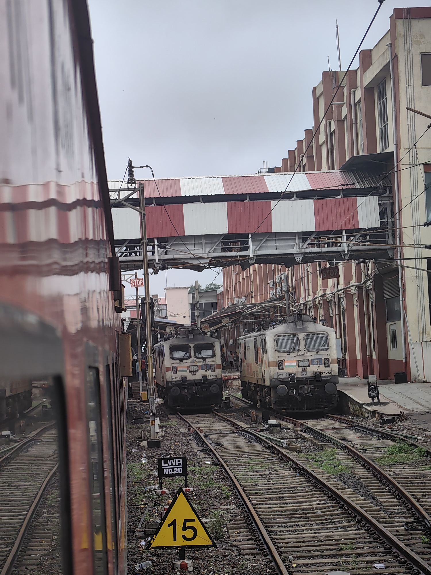 The image captures a scene at the Bhopal Junction railway station in Bhopal, India. In the foreground, the side of a red and white striped train carriage is visible, reflecting the surrounding environment. Two electric locomotives, identified as WAP 7 and bearing the number 37256, are positioned on parallel railway tracks. The locomotives are cream-colored with red and brown accents and feature the Indian flag emblem.
Overhead, a pedestrian bridge with alternating red and white striped awnings spans the tracks, connecting buildings on either side. The buildings themselves are a mix of architectural styles, with one prominently featuring red columns and cream-colored walls, interspersed with windows. Electrical wires and support structures for the overhead power supply for the trains are also visible.
On the ground, numerous railway tracks are present, with ballast and scattered debris. A yellow triangular sign with the number "15" in black is mounted on a pole, indicating a speed limit. Another black and white rectangular sign reads "LWR NO.20". A red "STOP" sign is also visible on a pole. The sky is overcast and grey. The overall atmosphere suggests a busy railway station environment.