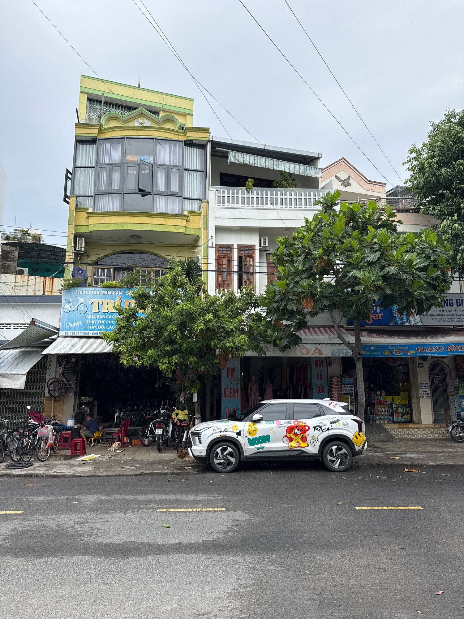The image shows a street scene in Nha Trang, Vietnam. On the left is a multi-story building painted yellow and green, with a prominent bay window on the second floor. Below, a bicycle shop is open, with bicycles displayed both inside and outside. A man is sitting on a red stool inside the shop. Two dogs are lying on the sidewalk in front of the shop. A white car decorated with stickers, including a cartoon character and smiley faces, is parked on the street. The car has the "Grab" logo on its side. In the background, other buildings are visible, including a white building with a balcony and a smaller building with a peaked roof. Trees line the street, and power lines crisscross the sky. The sky is overcast.