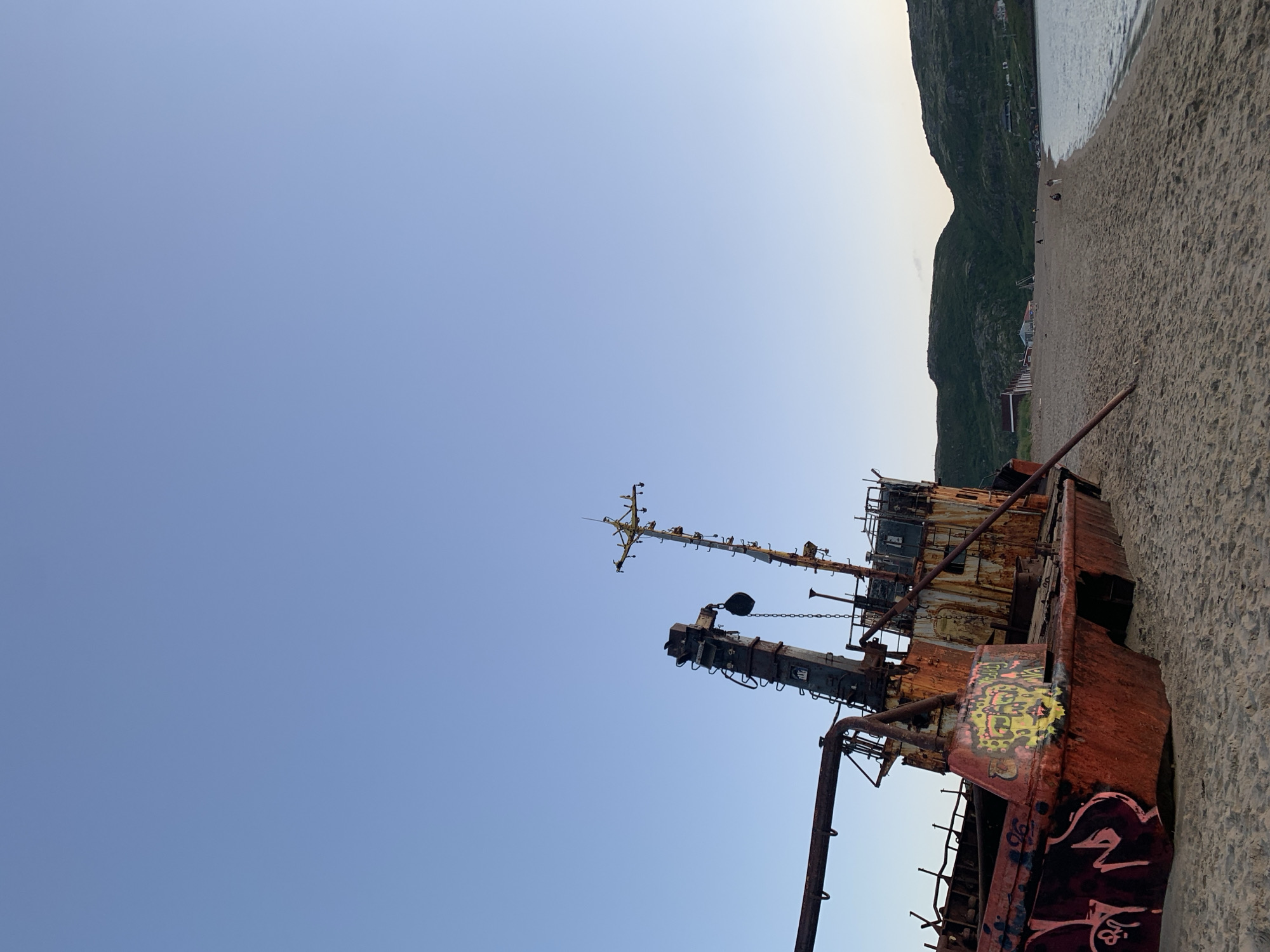 An abandoned, rusted shipwreck is beached on a sandy shore. The ship, adorned with graffiti, lies on its side. In the background, green, treeless hills rise above a calm body of water. A few small figures are visible walking on the beach and near some buildings nestled into the hillside. The sky is clear and a pale blue, indicating it is either early morning or late evening. This scene is located on the coast of Teriberka, Russia.