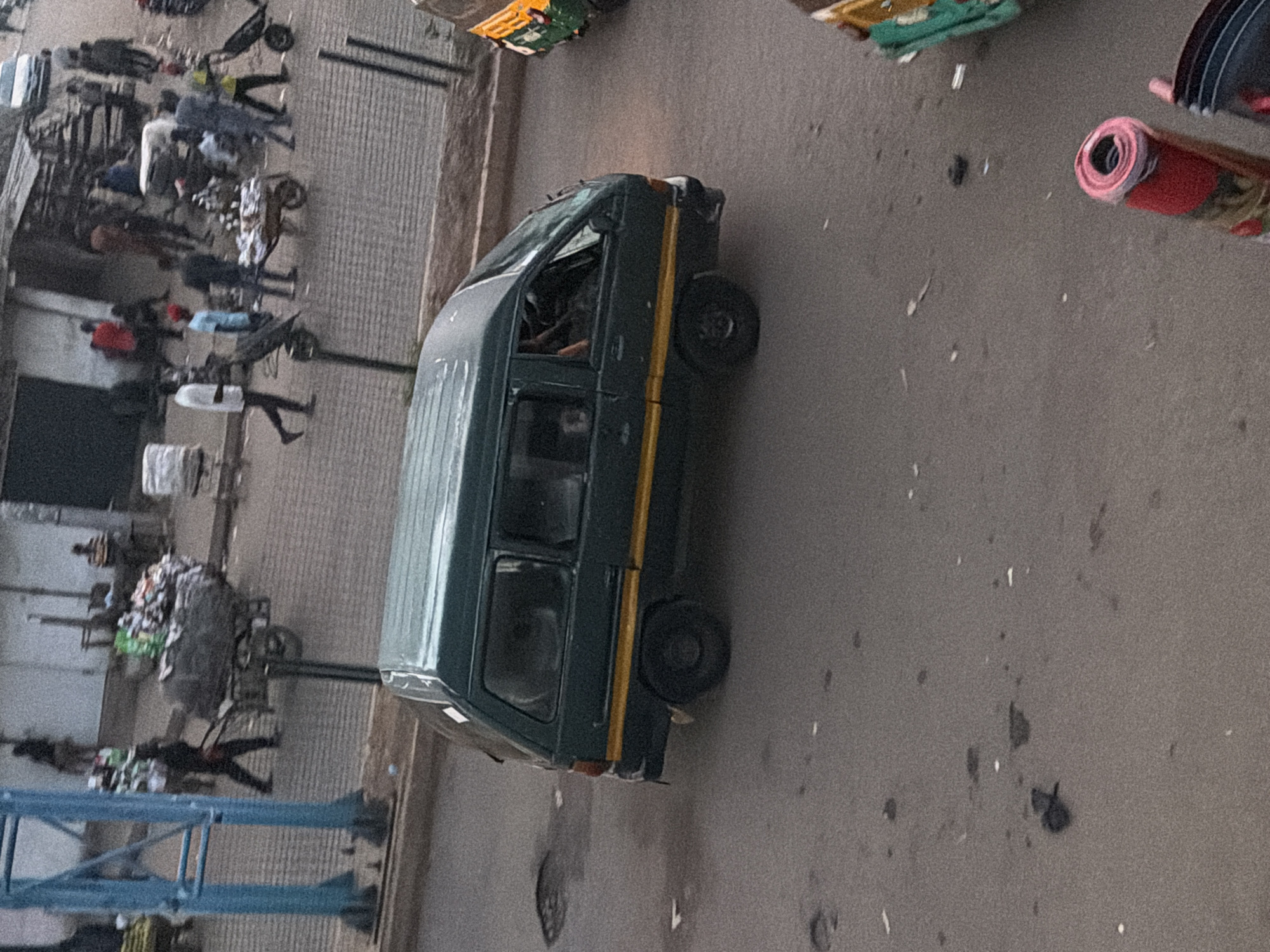 The image depicts a street scene in Bukuru, Nigeria, with a dark green minivan in the foreground. The minivan has a yellow stripe along its side and appears to be moving or parked on an asphalt road. The road has some puddles or uneven surfaces. In the background, there are several people, some carrying goods on their heads or using carts. There are also buildings and market stalls visible. A yellow and green auto-rickshaw is partially visible on the right side of the image. The overall impression is of a busy marketplace or street in a Nigerian city.