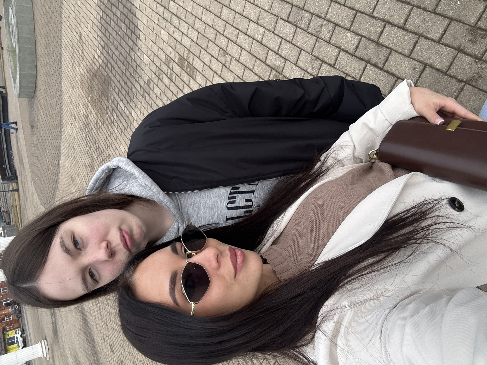 Two young women are lying side-by-side on a paved surface. The woman on the left has brown hair and is wearing a gray hooded sweatshirt and a black jacket. The woman on the right has long, dark hair, sunglasses, a beige sweater, and a white coat. She is holding a brown handbag. They are lying on a brick-paved walkway in Chaikovsky, Russia. A fountain is visible behind them. The photo appears to have been taken with a mobile phone.