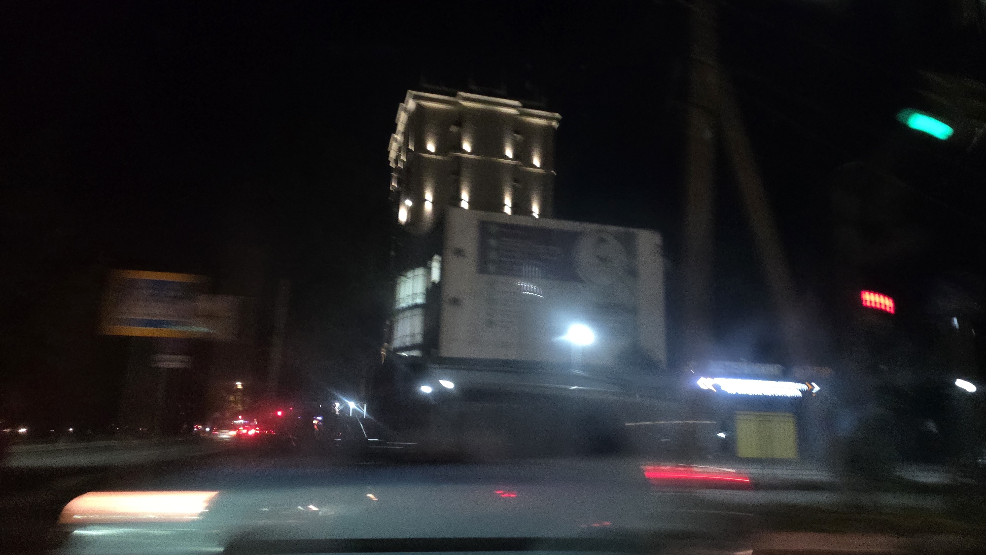 The image captures a nighttime street scene in Bishkek, Kyrgyzstan, characterized by motion blur indicating movement, likely from a vehicle. A tall, multi-story building with illuminated windows and distinct upward-facing lights is prominently featured. A large billboard or sign is attached to the front of this building. Further to the right, a traffic light with a green signal is visible, along with a red signal further down. A street sign with yellow and blue elements is present on the left side of the frame. Red taillights of vehicles and streaks of light suggest ongoing traffic. The environment appears to be an urban area with street lighting and infrastructure.