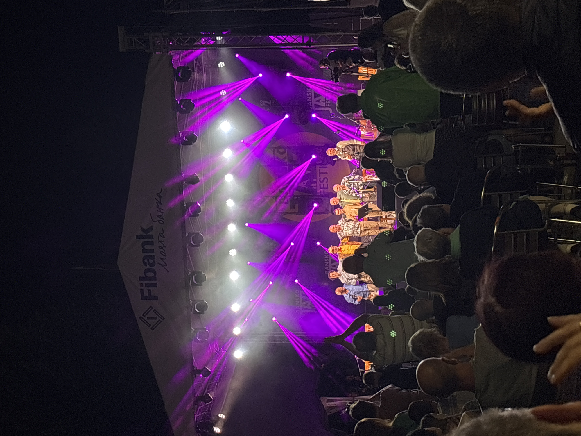 An outdoor nighttime music festival is depicted, with a stage adorned with purple lights. The backdrop of the stage features the "Fibank" logo and "Bansko Jazz Festival" text. A group of musicians, predominantly older men, are on stage, some holding instruments, and they appear to be interacting with each other. The audience, seated and standing, is facing the stage, with many people holding up their phones, presumably to record or photograph the event. The overall atmosphere suggests a lively and engaging musical performance taking place in the city of Bansko, Bulgaria.