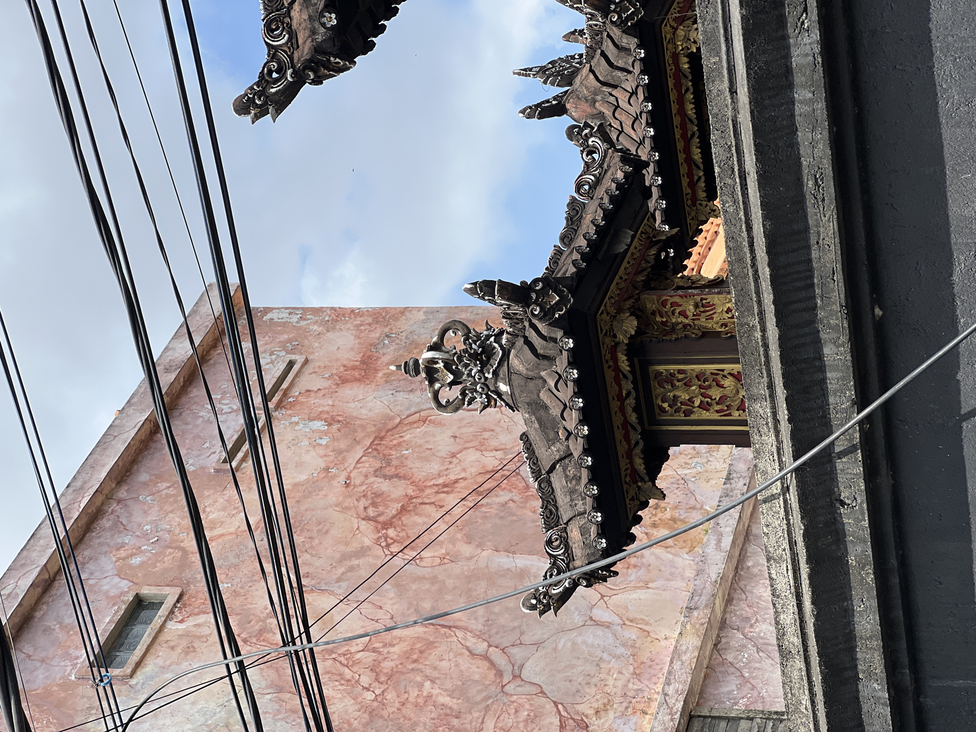 Looking up, the photo shows a building with a reddish-pink marbled facade, intersected by black electrical wires. Attached to the building is an ornately carved, dark-wood structure, decorated with small white floral details and some gold paint underneath. The sky is visible in the upper portion of the frame and is light blue with some clouds. The right side of the frame shows a textured, dark gray structure. The image was taken in North Kuta, Indonesia.