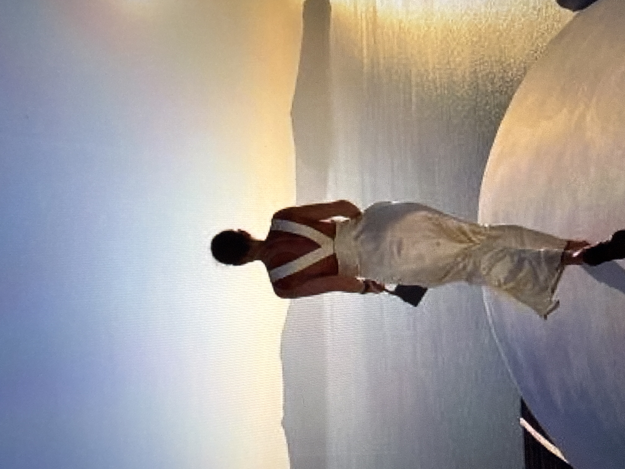 A woman is standing on a circular platform, facing away from the camera, looking out at a body of water and mountains in the distance. The sun is setting, casting a warm glow across the sky. The woman is wearing a long, flowing white dress and carrying a small black purse. The platform appears to be on a rooftop or balcony, with a railing visible in the foreground. The overall scene is serene and picturesque. The specific location in Găvănoasa, Moldova is not discernible from the image.