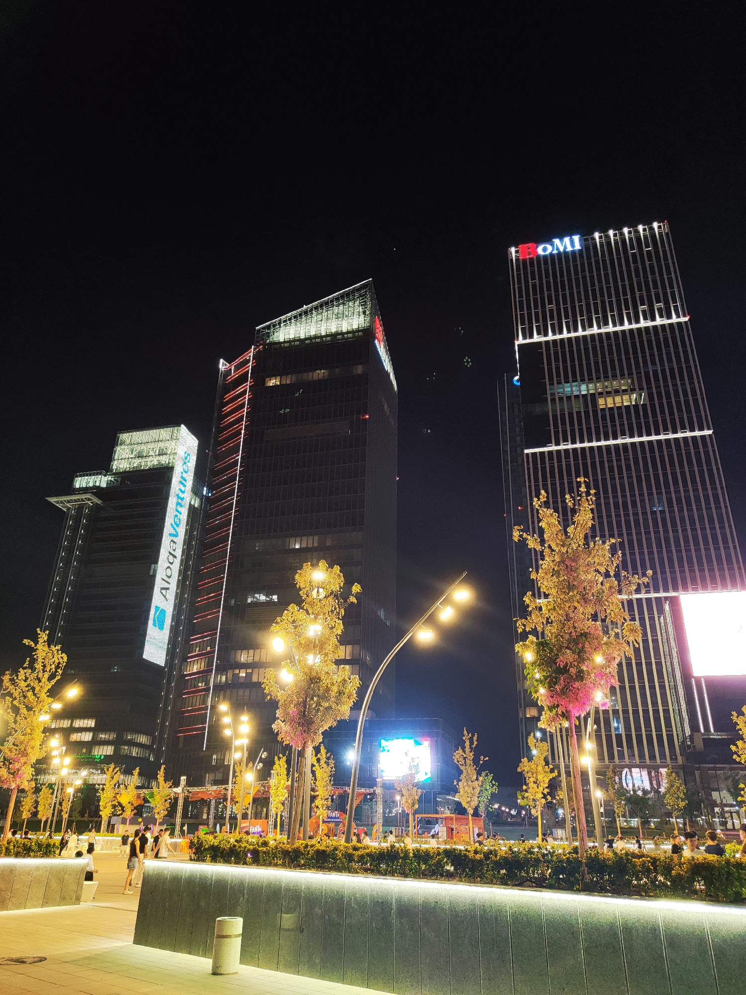 This is a nighttime exterior shot of the modern business district of Tashkent, Uzbekistan, specifically the IQ-Tower, also known as the "Oriyat Don-Don" complex, and the adjacent Aloqa Ventures building. The IQ-Tower, on the right, prominently displays the red and blue "OMI" logo at its apex, with vertical white lights illuminating its facade. The Aloqa Ventures building, on the left, features a large, vertical illuminated sign with white text on a blue background. Both skyscrapers are tall and modern, with glass and metal exteriors.
In the foreground, a well-lit public space with manicured hedges and strategically placed trees adorned with pink and yellow lights creates a welcoming atmosphere. Several streetlights with multiple bulbs illuminate the area. A few people are visible walking and socializing in this space, dressed in casual attire. The overall scene depicts a vibrant and illuminated urban environment, showcasing the city's contemporary architecture and lively public spaces at night.