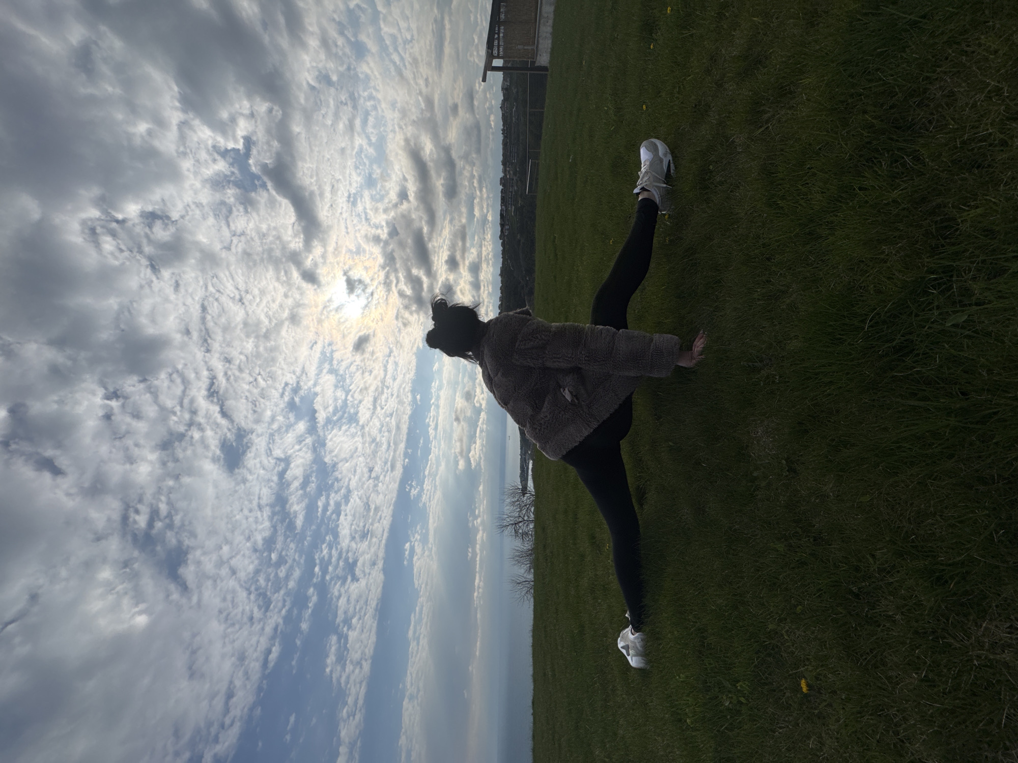 A woman with dark hair pulled into a bun stands on a grassy field with her legs spread wide, one foot forward and one foot back. She wears black leggings, white sneakers, and a tan, fuzzy jacket. The sky is filled with puffy white clouds, and the sun is obscured by the clouds. In the background, a building is partially visible. The photo appears to have been taken in Topola, Bulgaria, but no identifying landmark is visible.