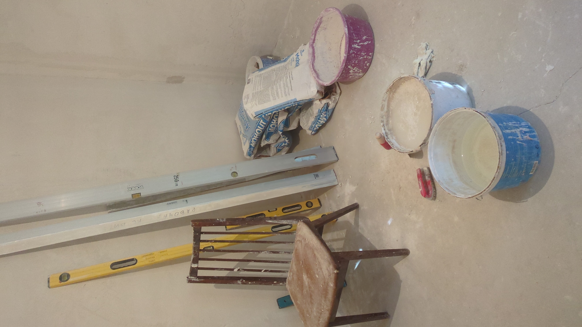 The image depicts an indoor scene, likely a room under renovation or construction, with building materials scattered on the floor. The floor is concrete and appears dusty.
In the foreground, two buckets are visible. The larger one, on the right, is blue and white, containing clear liquid and some white sediment at the bottom. The smaller bucket, slightly to the left and behind, is white and appears to contain a mixture, possibly plaster or mortar, with some bubbles on the surface. A small red and black tool, possibly a trowel or brush, lies between these two buckets. To the right of the white bucket, a pair of light-colored gloves are partially visible.
Dominating the mid-ground are several bags of building material, white with blue lettering that appears to spell "MOHOUT." These bags are stacked leaning against the wall. To the left of these bags, leaning against the wall, are three long, flat leveling tools or rulers. One is yellow, and the other two are silver/gray. The yellow one has black markings and a bubble level. The silver/gray ones also have markings and bubble levels.
In the left mid-ground, a wooden chair with a slightly worn, dusty seat and a slatted back is positioned. A small, blue object, possibly a piece of tile or a tool, is visible on the floor near the chair's legs.
The walls are a light, neutral color, appearing to be plastered or painted. The overall impression is one of a work in progress, with materials ready for use.
The scene is devoid of people. No specific geographical markers for Chișinău, Moldova are present within the image itself.