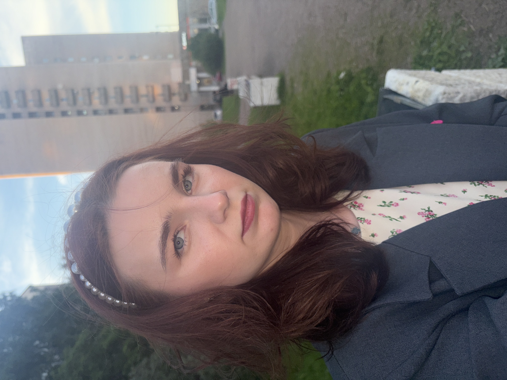 A young woman is captured in a close-up portrait, looking directly at the camera with a calm and direct gaze. She has fair skin, long, wavy dark reddish-brown hair, and light blue-green eyes. Her lips are subtly tinted with a rosy color. A white pearl-studded headband adorns the left side of her head, nestled in her hair. She is wearing a dark, possibly navy or charcoal, blazer over a light-colored top featuring a delicate floral pattern of small pink and green blossoms.
The setting appears to be an urban outdoor environment, likely in late afternoon or early evening, characterized by soft, diffused natural light. In the background, a large, multi-story building with a neutral, light-colored facade is visible, suggesting an institutional or older architectural style common in cities. Beyond the building, a light blue sky is discernible. To the woman's right, there is green foliage and a glimpse of what appears to be a light-colored stone or concrete structure, possibly a bench or wall. The background is softly blurred, keeping the focus on the subject. There is no visible text in the image. The scene is consistent with an urban landscape such as Saint Petersburg, Russia.