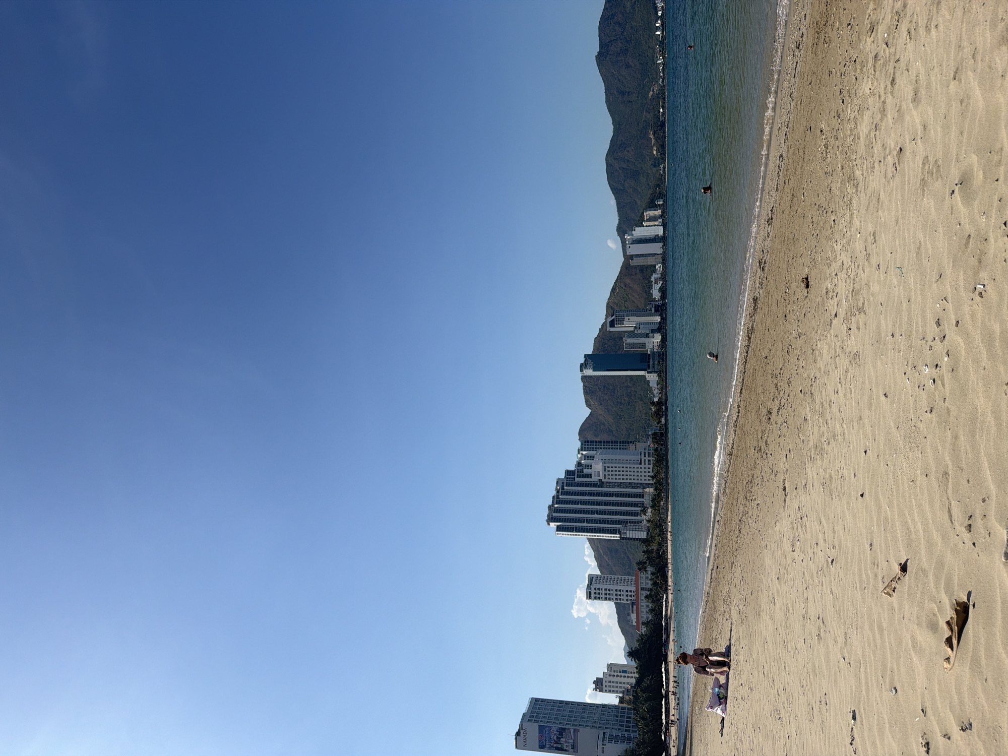The image depicts a beach scene in Bac Nha Trang Ward, Vietnam. The view is oriented such that the sky occupies the left half of the frame, transitioning to a beach on the right. The sea borders the beach, with some people visible in the water. Mountains form a backdrop along the coastline, punctuated by high-rise buildings. Near the bottom of the frame, a person is sitting on the beach. The sand shows ripples and scattered debris.