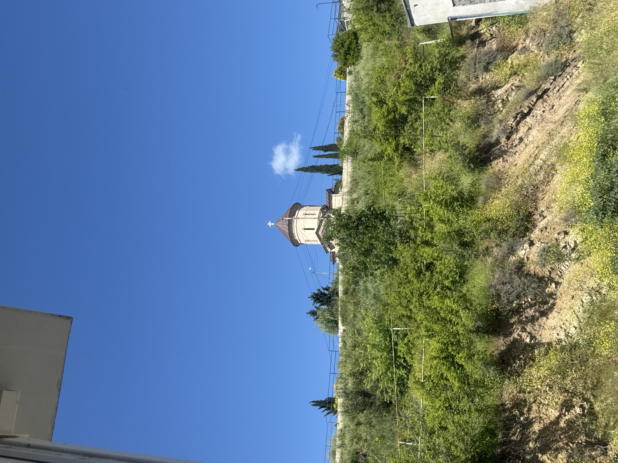 The image depicts a clear, bright blue sky dominating the upper portion, with a building visible in the lower left corner. An Orthodox church stands out against the sky, its architectural details including a cross on top of a rounded tower. Power lines run parallel to the church towards the upper right corner of the image. The church is built on a hillside covered with green vegetation, bushes, and trees that fill the right side of the frame. A dirt embankment is also visible. The location is likely Tbilisi, Georgia, given the style of the church.