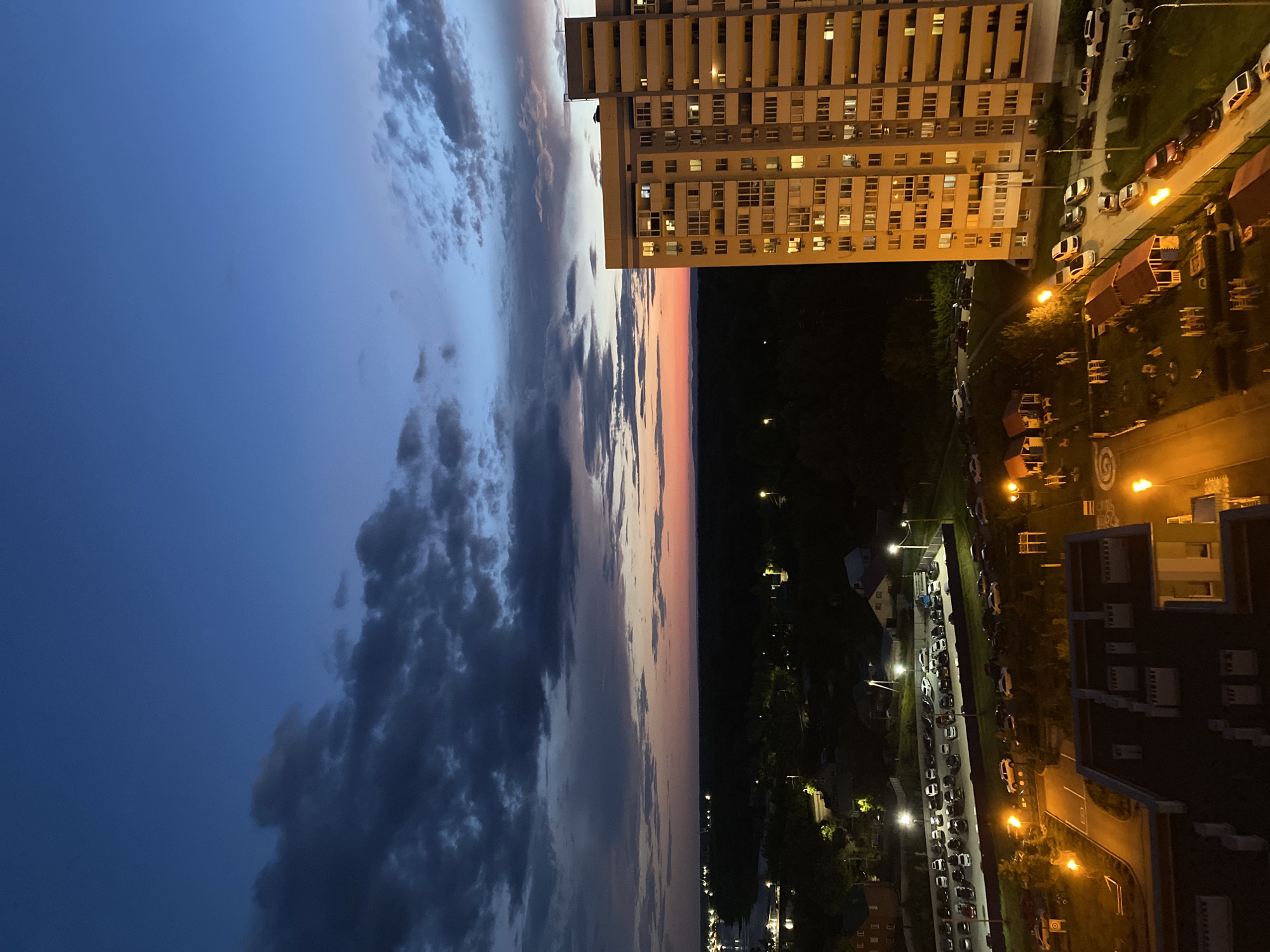 The image depicts a twilight scene over a city, likely Novokuybyshevsk, Russia. The sky transitions from a deep blue at the top to lighter hues of pink and orange near the horizon, with dark clouds scattered across the upper portion.
Dominating the right side of the frame is a tall, multi-story apartment building with numerous lit windows, indicating occupancy. In the foreground and midground, there are residential buildings, some with visible roof structures and windows. A paved area with parked cars is visible, along with streetlights casting a warm glow on the asphalt. Further into the scene, a dense area of trees forms a dark line against the fading light, suggesting a park or wooded area. Distant lights twinkle on the horizon, hinting at the spread of the city. The overall atmosphere is one of a quiet evening in an urban residential area. There are no people or discernible actions in the image.