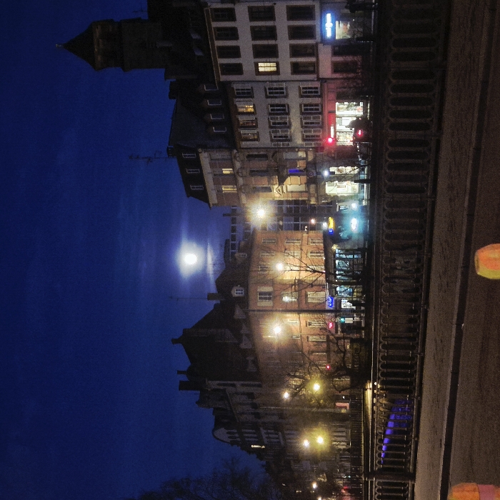 The image captures a serene nighttime cityscape, likely along a canal or river, under a deep blue sky illuminated by a prominent full moon.
On the left side of the frame, a dark, imposing building featuring a pointed tower, possibly a church or a historic clock tower, stands tall against the night sky. To its right, a row of multi-story buildings with traditional European architecture lines the street or embankment. Many windows of these buildings are lit with warm light, suggesting ongoing activity or residences within.
At street level, various commercial establishments are visible, marked by glowing signs in vibrant colors such as blue, red, and white. While the specific text on these signs is indistinct due to the distance and lighting, they contribute to the lively urban atmosphere. Warm-toned streetlights cast a yellow-orange glow, illuminating the facades of the buildings and parts of the street below. Sparse, bare tree branches are visible among the buildings and streetlights, suggesting a colder season like winter or late autumn.
To the right, a dark, decorative metal railing, possibly part of a bridge or an elevated walkway, extends horizontally across the midground. Beyond this railing, a dark surface, presumably a street or pathway, occupies the foreground. Reflections of the artificial city lights are discernible, particularly on the lower right, hinting at the presence of water, such as a canal or river, running parallel to the buildings.
The sky is a rich, dark blue, with subtle cloud formations. The most striking natural feature is the bright, full moon, positioned prominently in the upper-mid section of the sky, casting a gentle, ethereal glow over the scene. The overall impression is one of a peaceful yet active urban night, where the natural moonlight harmonizes with the artificial luminescence of the city. The time of day is clearly night. No specific individuals or discernible human activities are clearly visible.