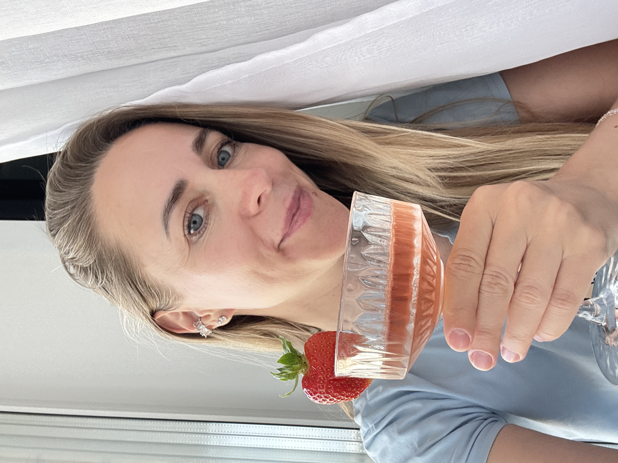 A blonde woman with blue eyes holds a champagne coupe filled with a pink beverage and decorated with a strawberry. She is wearing a light blue t-shirt and a silver ear cuff. The image was taken indoors, with natural light illuminating the scene from the left. A white curtain hangs on the right side of the frame. The woman is looking towards the camera and smiling. The setting is a residential environment, likely an apartment, in the city of Chaikovsky, Russia.